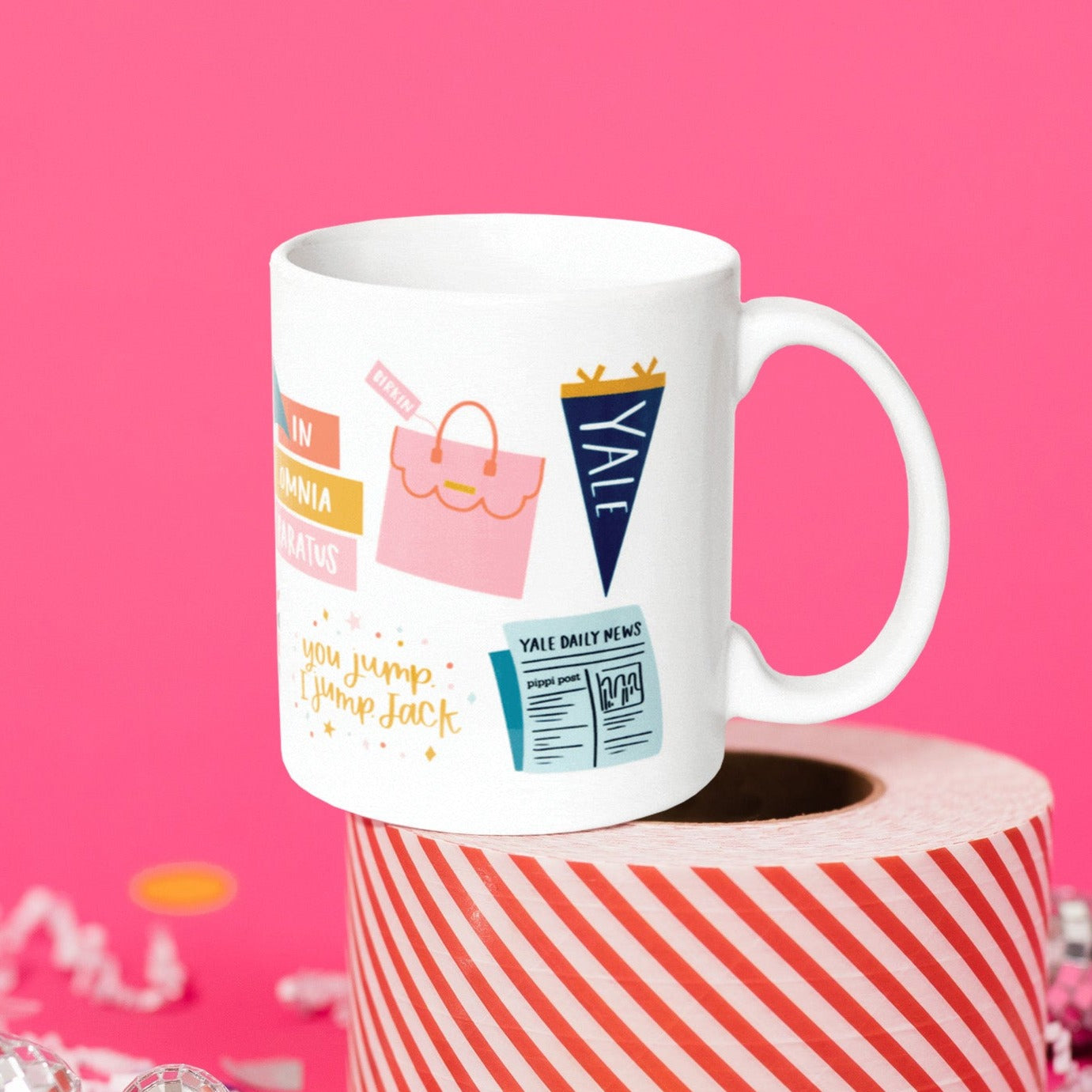 On a hot pink background sits a mug atop a red and white striped roll of packing tape with white crinkle and big, colorful confetti scattered around. There are mini disco balls. This Gilmore Girls inspired white mug says colorful illustrations from the show. There is a Birkin pink purse, a navy school banner that says “YALE”, a "YALE DAILY NEW" newspaper, and colorful stars and dots. It says "you jump I jump jack.”