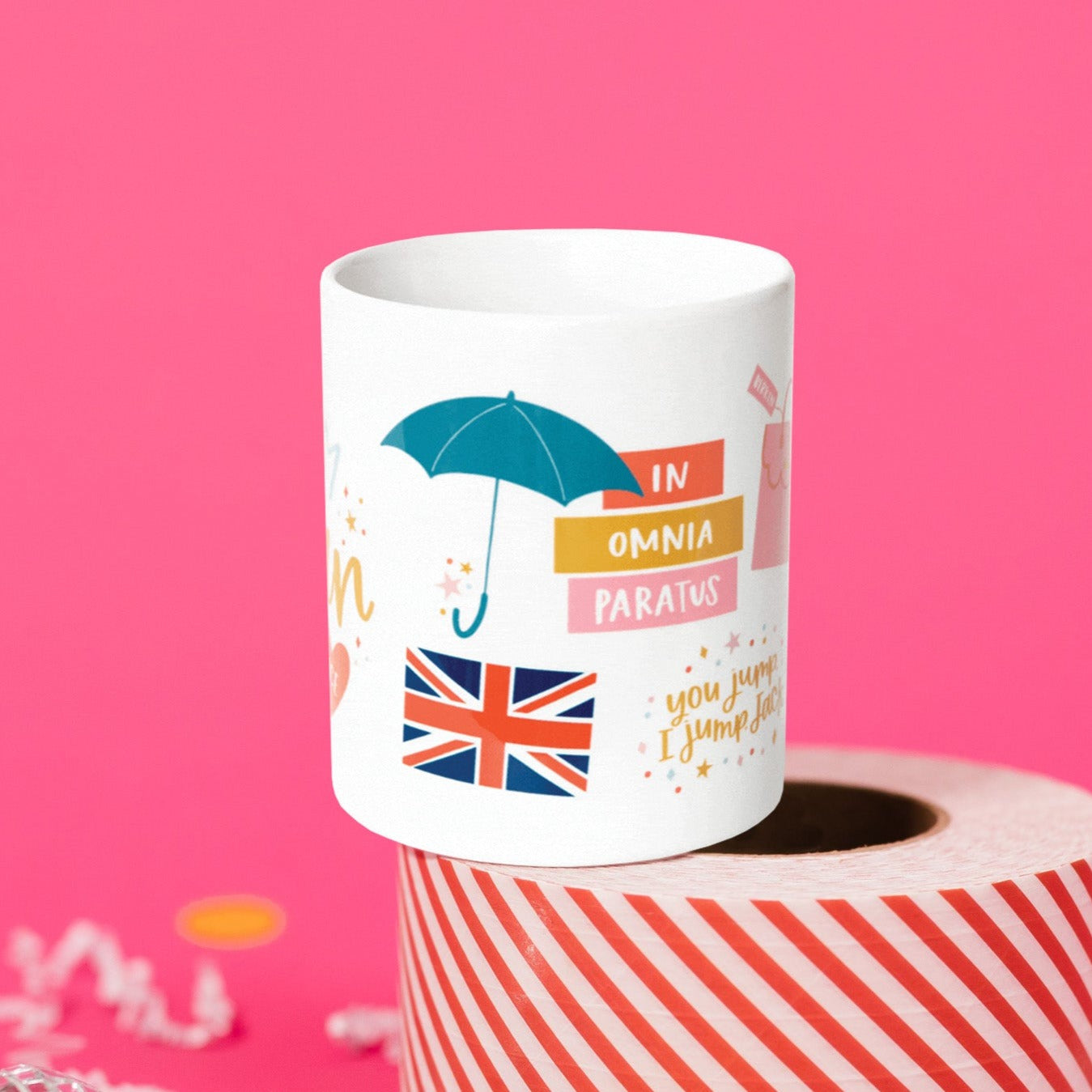 On a hot pink background sits a mug atop a red and white striped roll of packing tape with white crinkle and big, colorful confetti scattered around. There are mini disco balls. This Gilmore Girls inspired white mug has colorful illustrations. It has a teal umbrella, a Great Britain flag, colorful stars and dots and it says "you jump I jump jack.” There are three colorful rectangles. It says “IN”, "OMNIA" and “PARATUS.”