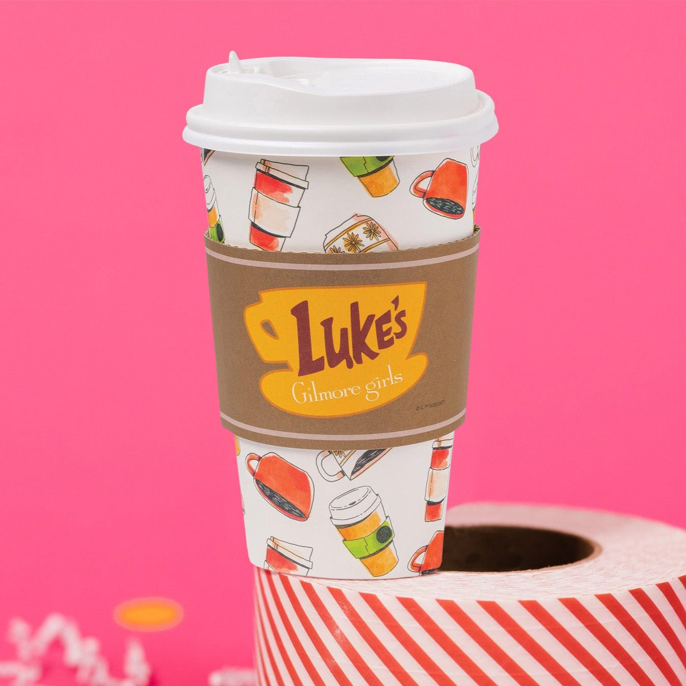 On a hot pink background sits a cup atop a red and white striped roll of packing tape with white crinkle and big, colorful confetti scattered around. This Gilmore Girls inspired white to-go coffee cup has a white lid and colorful illustrations of coffee mugs and to-go cups all over with a kraft band around the center that has a sunny mustard illustration of a mug and the words "Luke's Gilmore girls" on it.