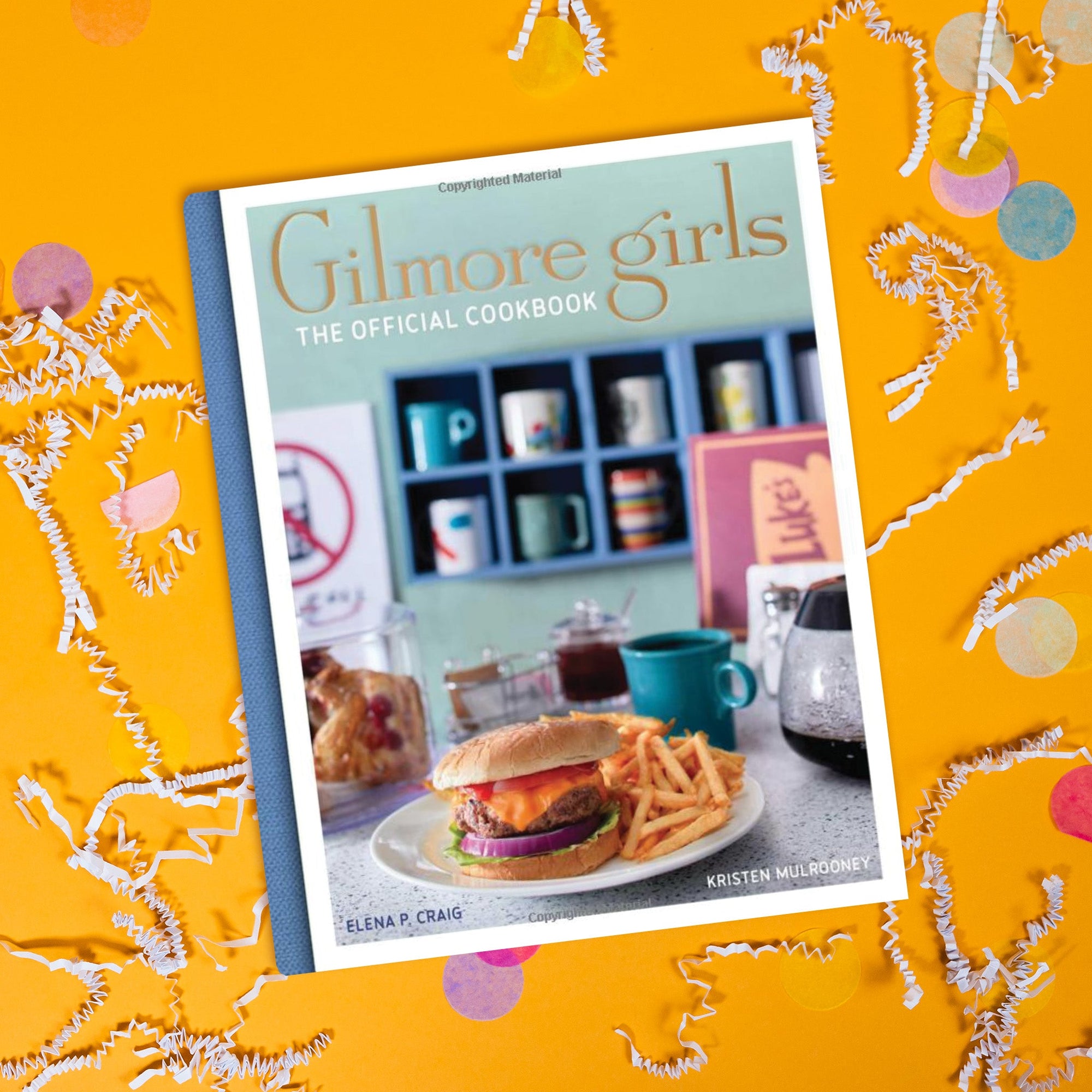 On a sunny mustard background sits a book. This Gilmore Girls inspired cookbook says in gold serif font "Gilmore girls" and in all caps, white block font "The Official Cookbook." It has a picture of a diner in the background with a plate of hamburger and fries, with coffee and mugs.
