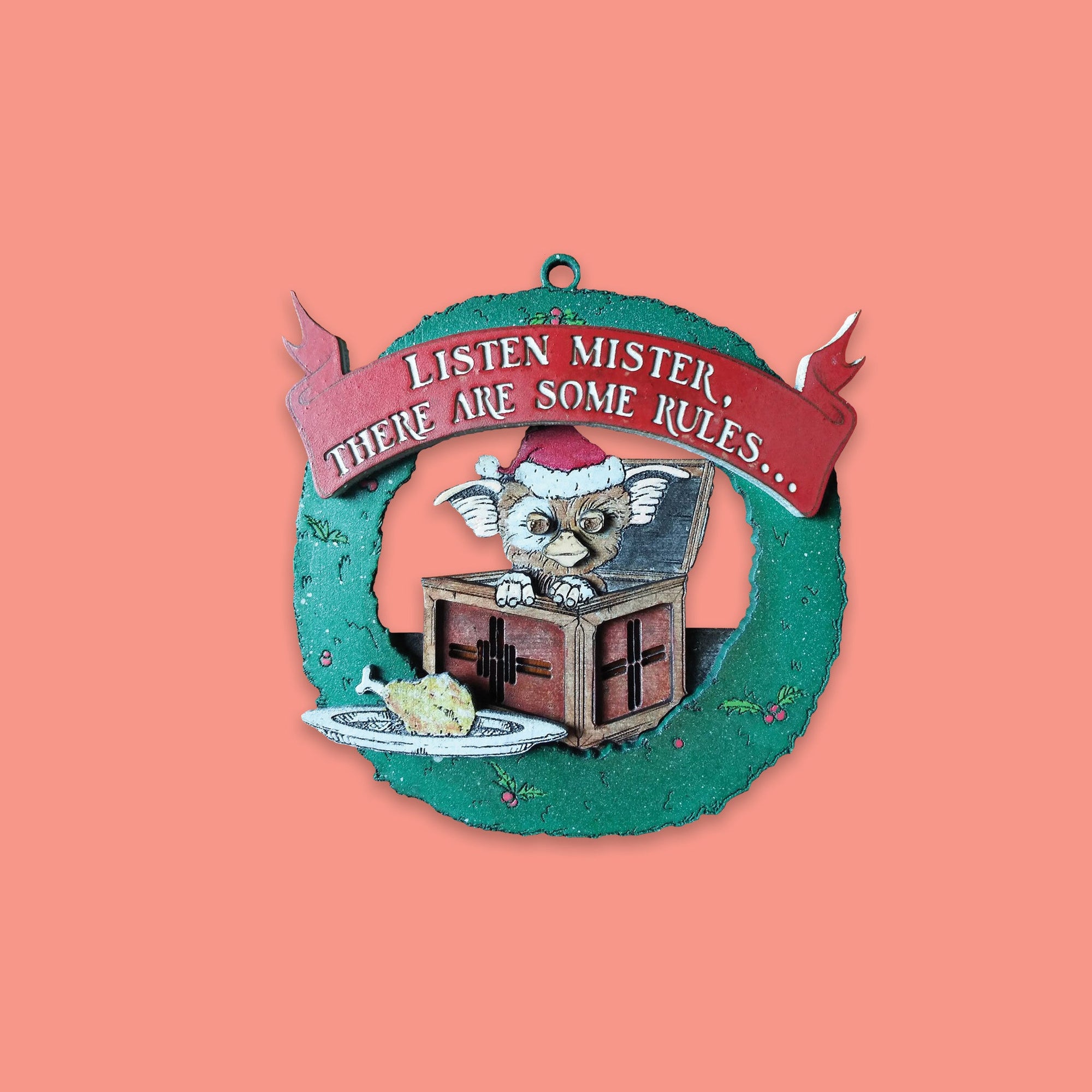 On a coral pink background sits a wreath ornament. This Gremlins inspired wood ornament is a green wreath with Gizmo wearing a santa hat and sitting in an opened box. There is a plate with a chicken leg on it. At the top is a red banner that says "LISTEN MISTER, THERE ARE SOME RULES..." in white lettering. 