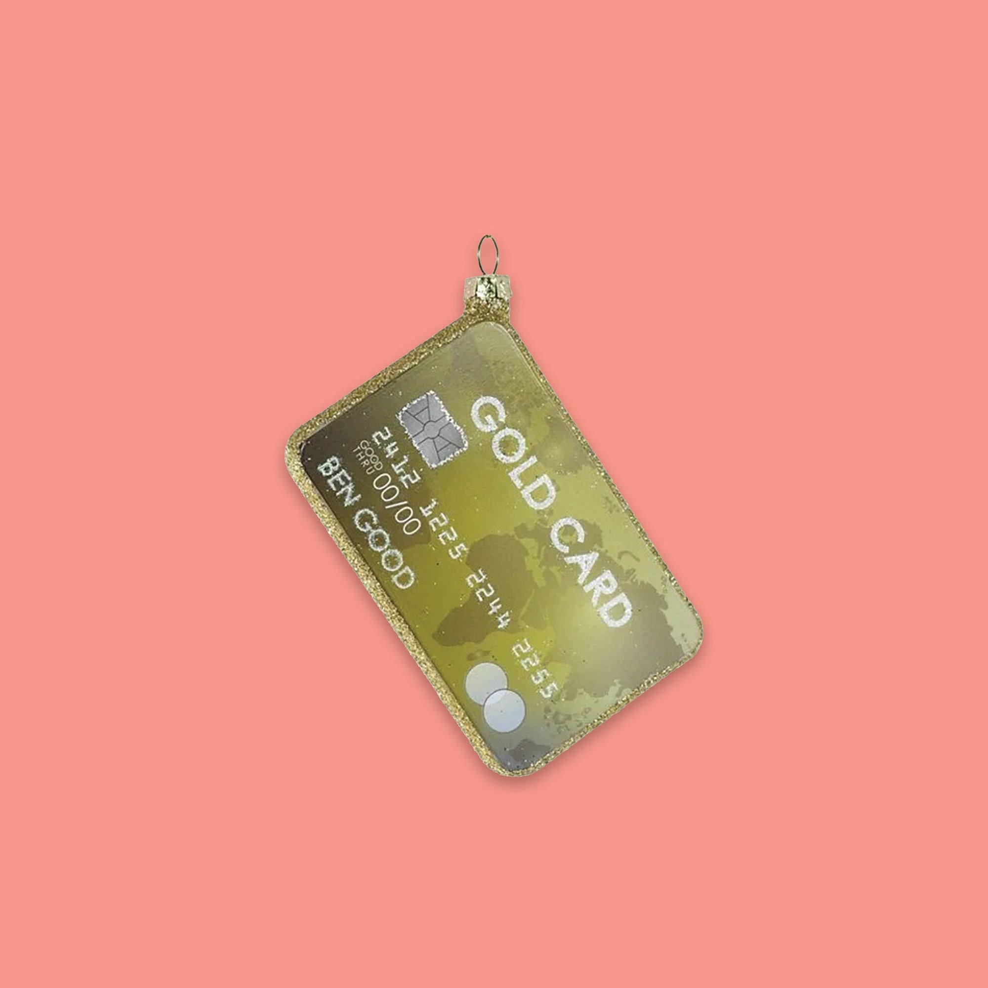 On a coral pink background sits an ornament. This is a glass ornament of a gold credit card. It says "GOLD CARD" and the name on it is "BEEN GOOD." It looks like a real credit card and has gold glitter around the edge.