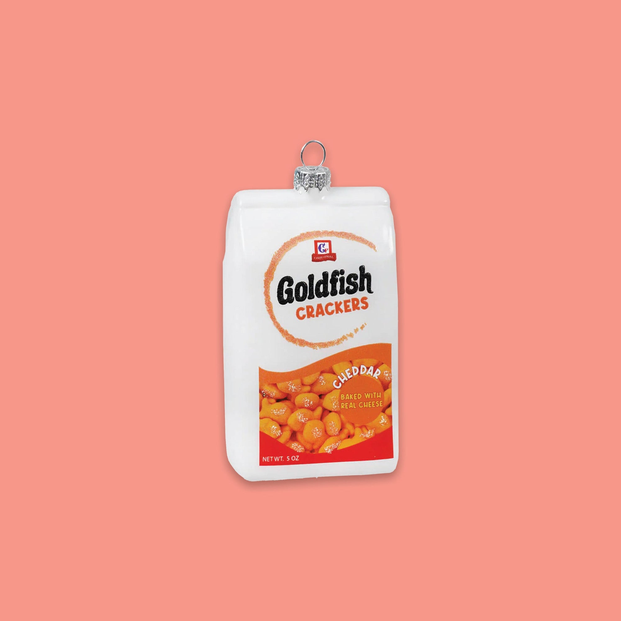 On a coral pink background sits an ornament. This is a white and orange bag of 'Goldfish Crackers' and there are goldfish on the front. It says "CHEDDAR BAKED WITH REAL CHEESE."