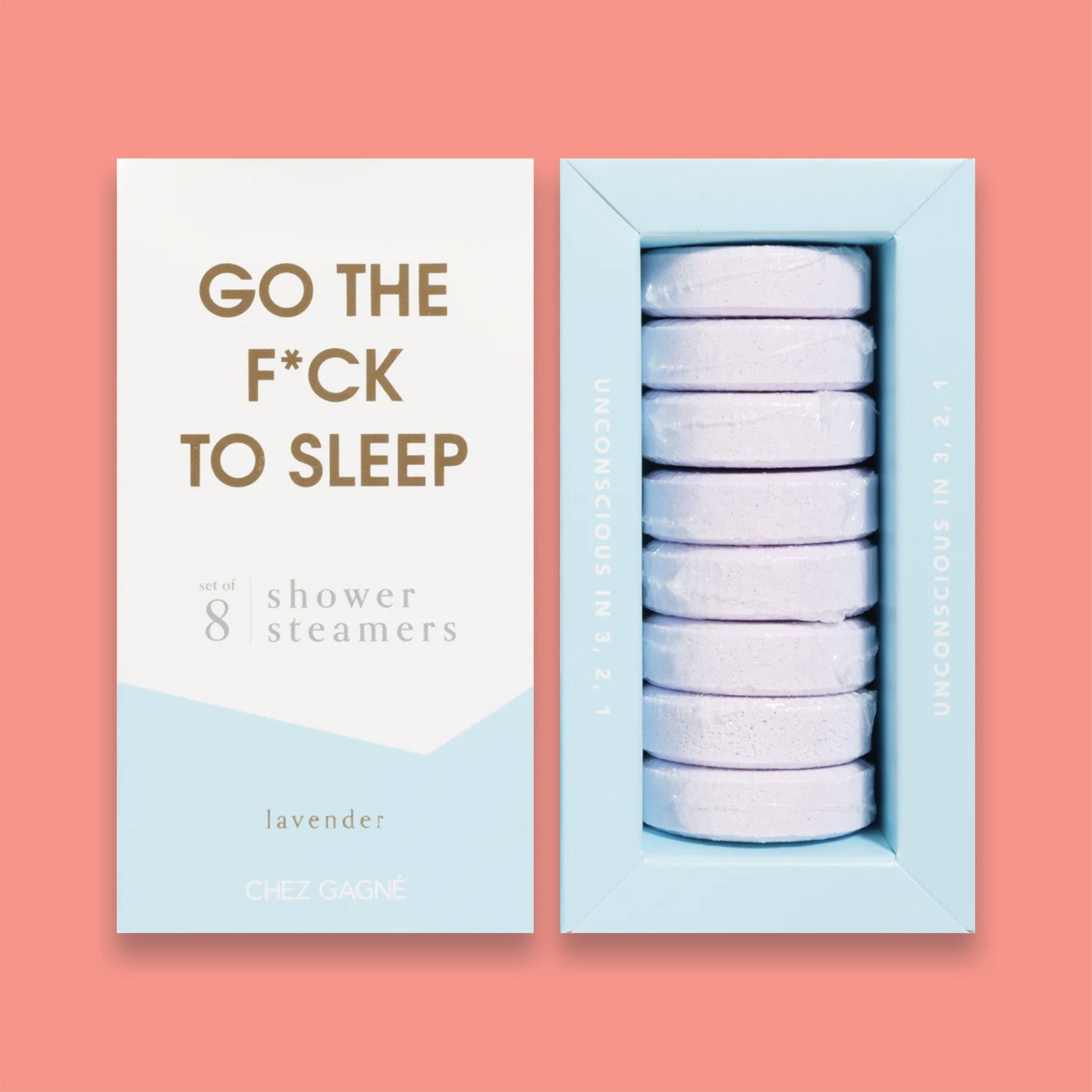 On a light coral background sits two boxes. This picture is a close-up of a white and light blue package that says "GO THE F*CK TO SLEEP" in gold foil, all caps block lettering. Under it says "set of 8" and " shower steamers" in grey, lowercase serif font. At the bottom it says "lavender" in gold foil, lower case serif font. To the right of it is a light blue box with light purple shower steamers in it. 