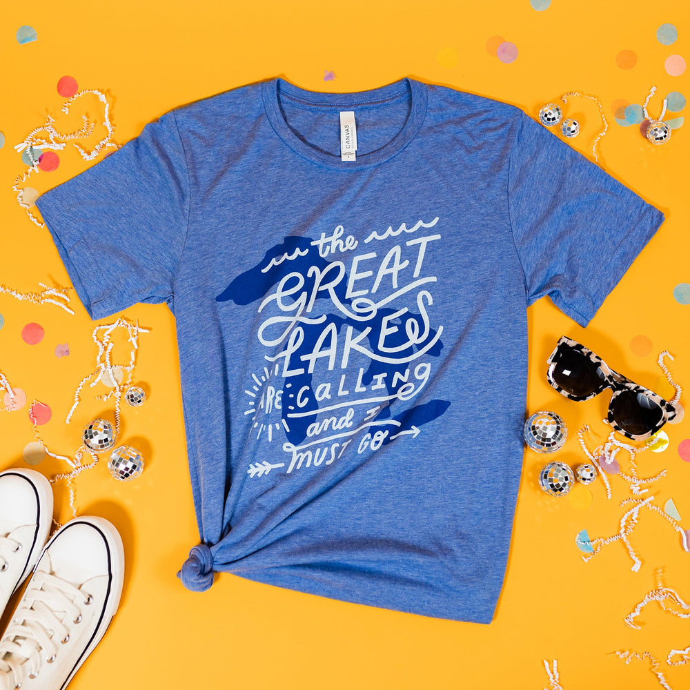 On a sunny mustard background sits a t-shirt with white crinkle and big, colorful confetti scattered around. There are mini disco balls, sunglasses, and a pair of white sneakers. This heather blue tee features an illustration of the Great Lakes printe in blue ink, with custom hand lettered script in white.