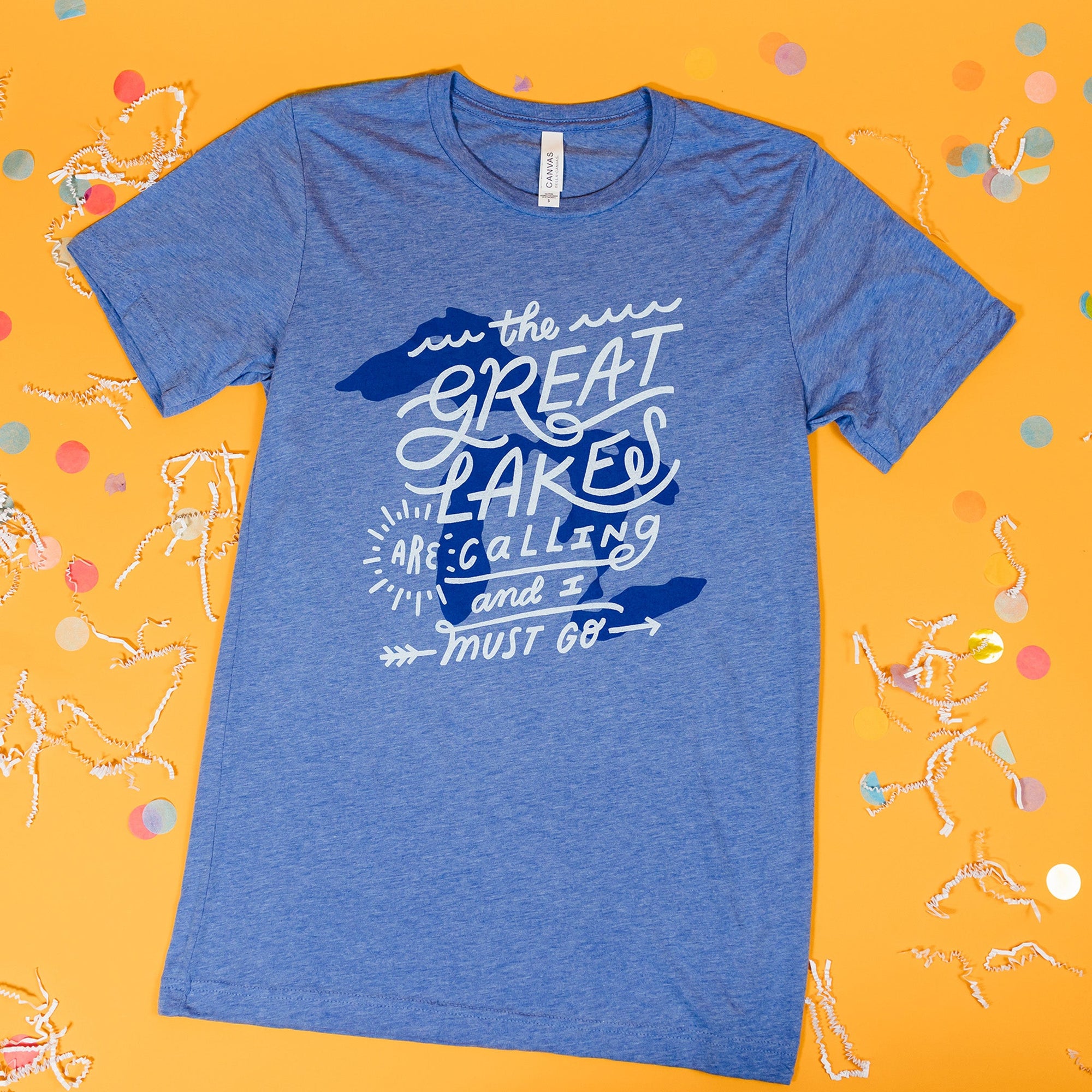 On a sunny mustard background sits a t-shirt with white crinkle and big, colorful confetti scattered around. This heather blue tee features an illustration of the Great Lakes printe in blue ink, with custom hand lettered script in white.