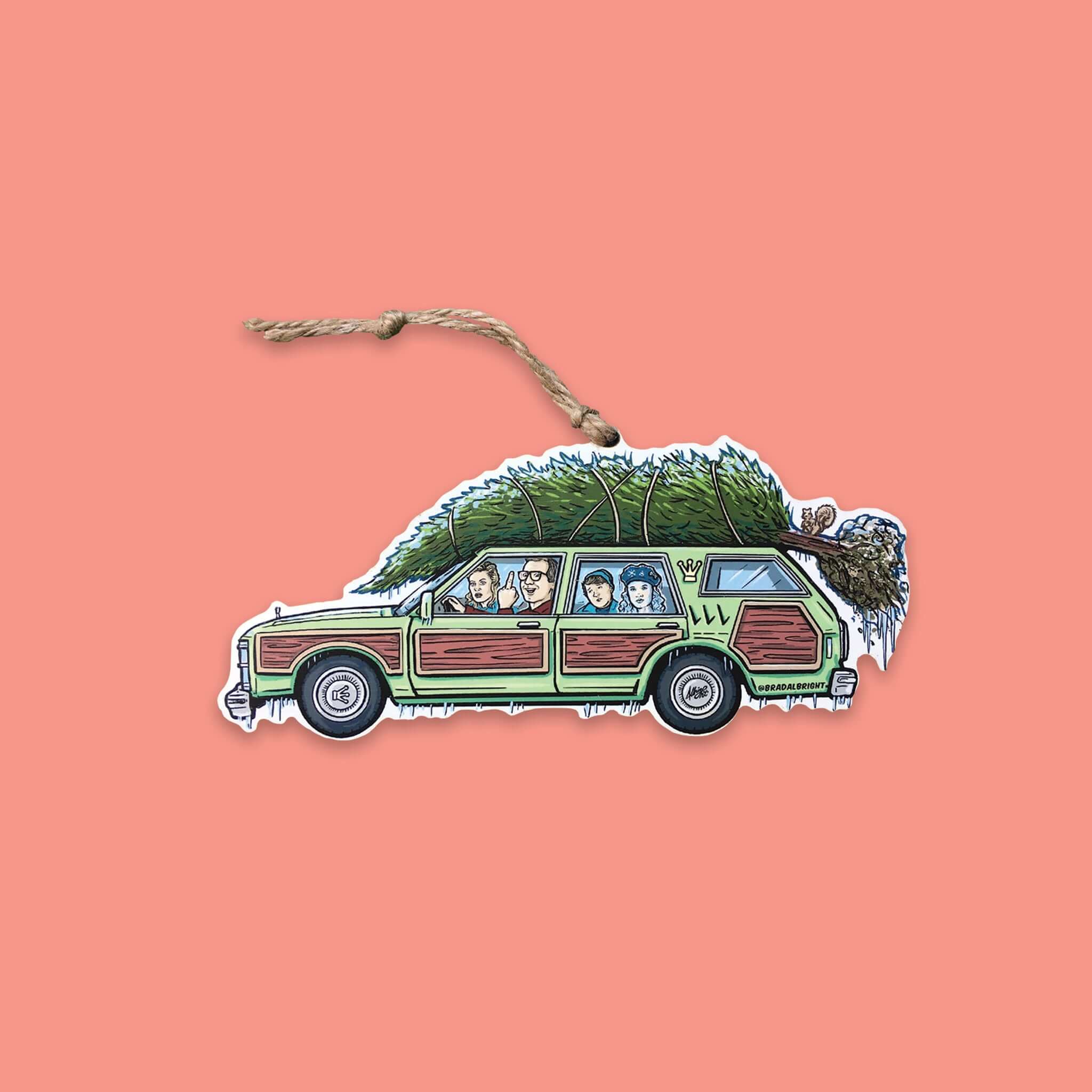 On a coral pink background sits an ornament. This Griswold Family Vacation themed wood ornament is of the family station wagon car with a christmas tree tied to the top. The Griswold family is sitting inside the car and Clark Griswold is giving the middle finger.