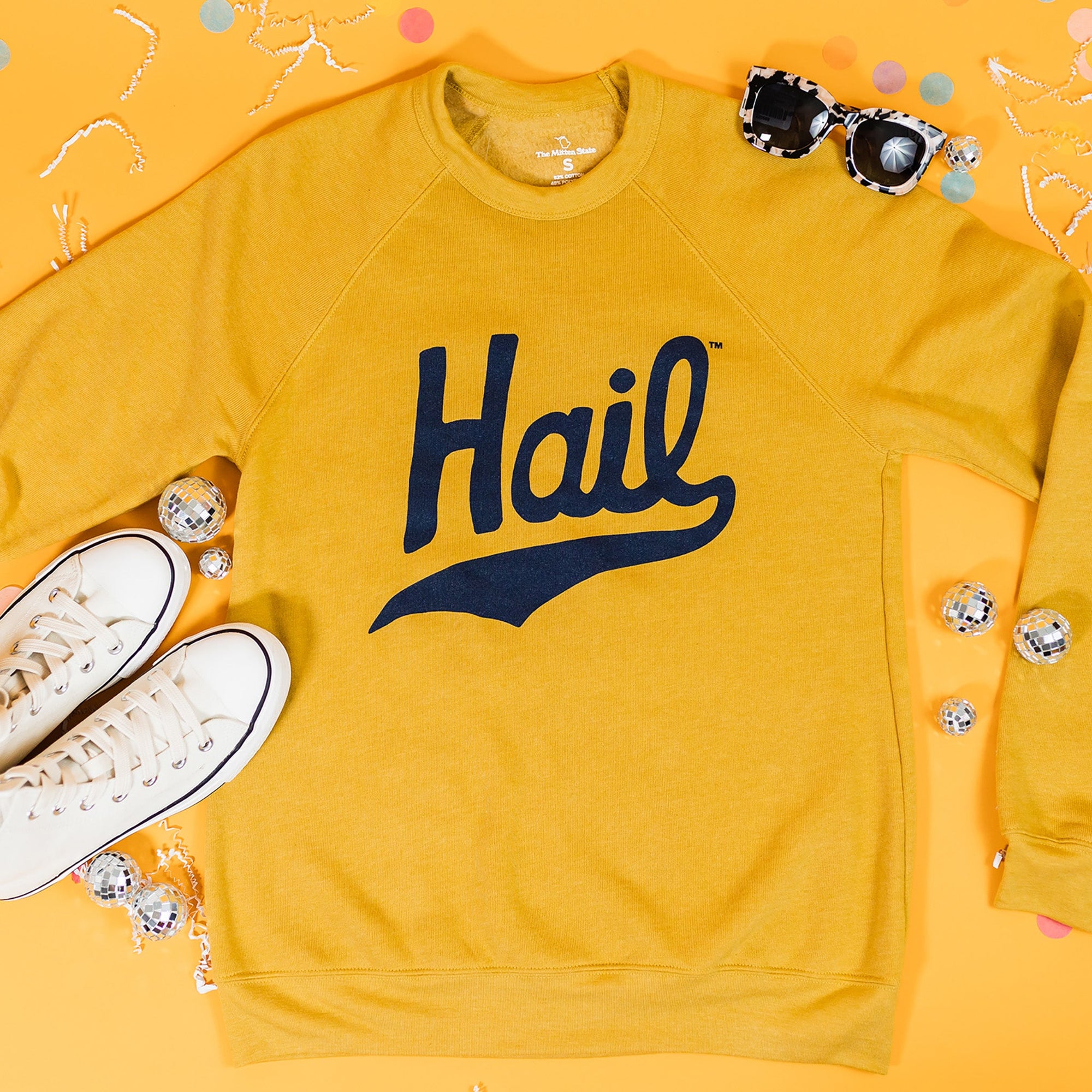 On a sunny mustard background sits a sweatshirt with white crinkle and big, colorful confetti scattered around. There are mini disco balls, sunglasses, and a pair of white sneakers. This mustard crewneck sweatshirt features a vintage, hand lettered "Hail" in navy.