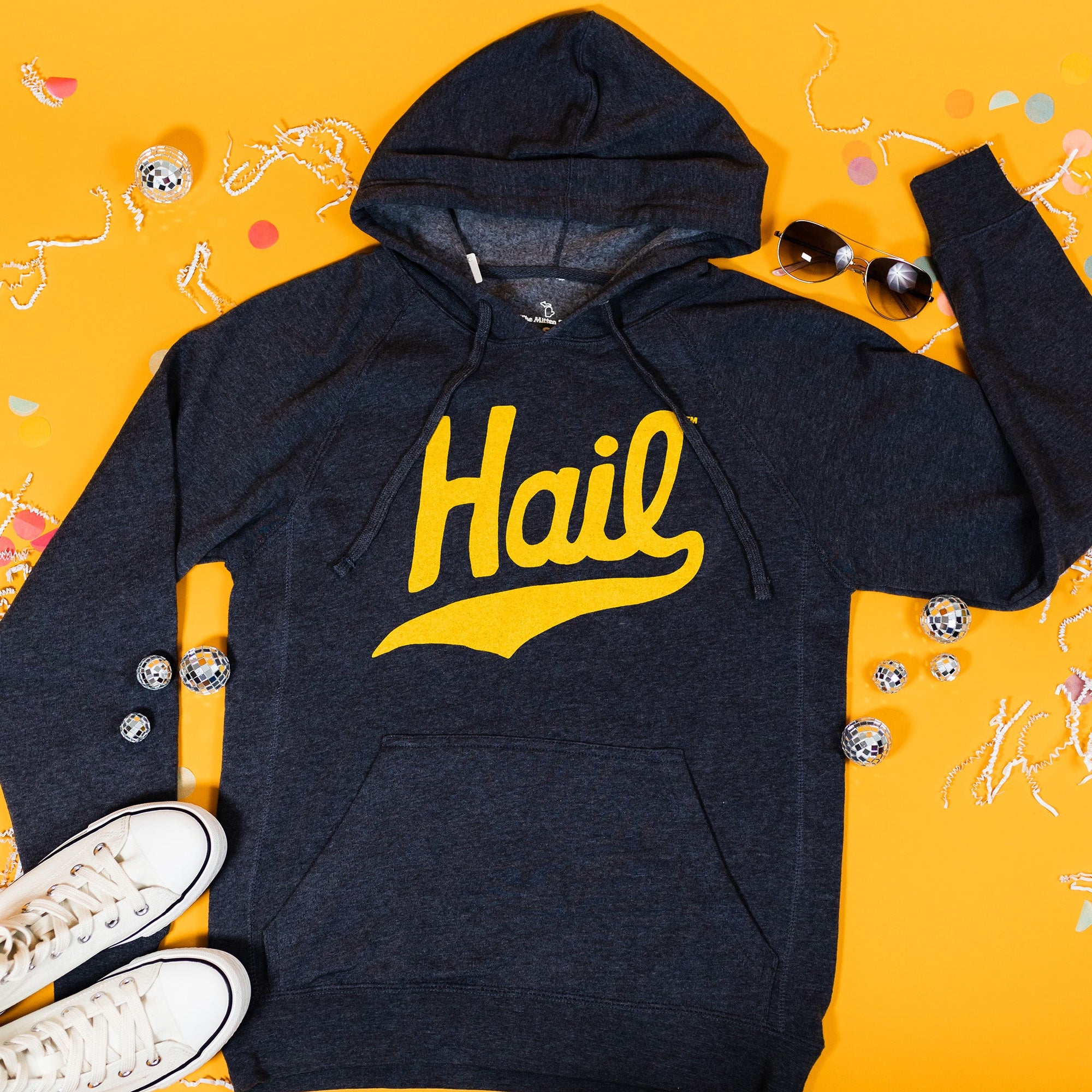 On a sunny mustard background sits a sweatshirt with white crinkle and big, colorful confetti scattered around. There are mini disco balls, sunglasses, and a pair of white sneakers. This navy sweatshirt features a vintage, hand lettered "Hail" in maize.