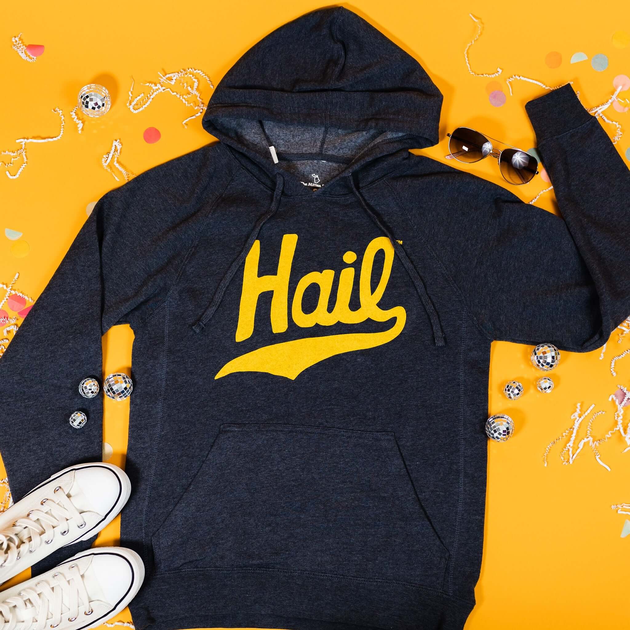 On a sunny mustard background sits a sweatshirt with white crinkle and big, colorful confetti scattered around. There are mini disco balls, sunglasses, and a pair of white sneakers. This navy sweatshirt features a vintage, hand lettered "Hail" in maize.