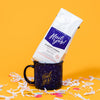 On a sunny mustard background sits a mug and a bag of coffee with white crinkle and big, colorful confetti scattered around. The mug is a navy blue campfire style mug with white splatter pattern. On the mug is a mustard yellow script font that says "Hail Yes!."  The white bag of coffee sits atop the mug. The coffee is Rock Paper Scissors 'Hail Yes!' in white hand lettering on a brilliant blue label. It is 'BLUEBERRY CRUMBLE GROUND COFFEE'. Net Wt. 12 oz. (340g)