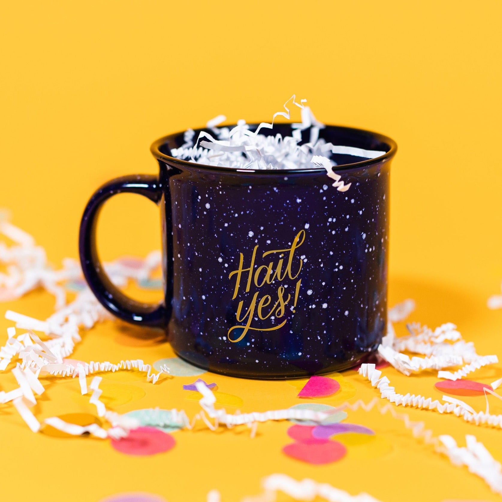 On a sunny mustard background sits a mug with white crinkle and big, colorful confetti scattered around. The mug is a navy blue campfire style mug with white splatter pattern. On the mug is a mustard yellow script font that says "Hail Yes!" and it is filled with white crinkle.