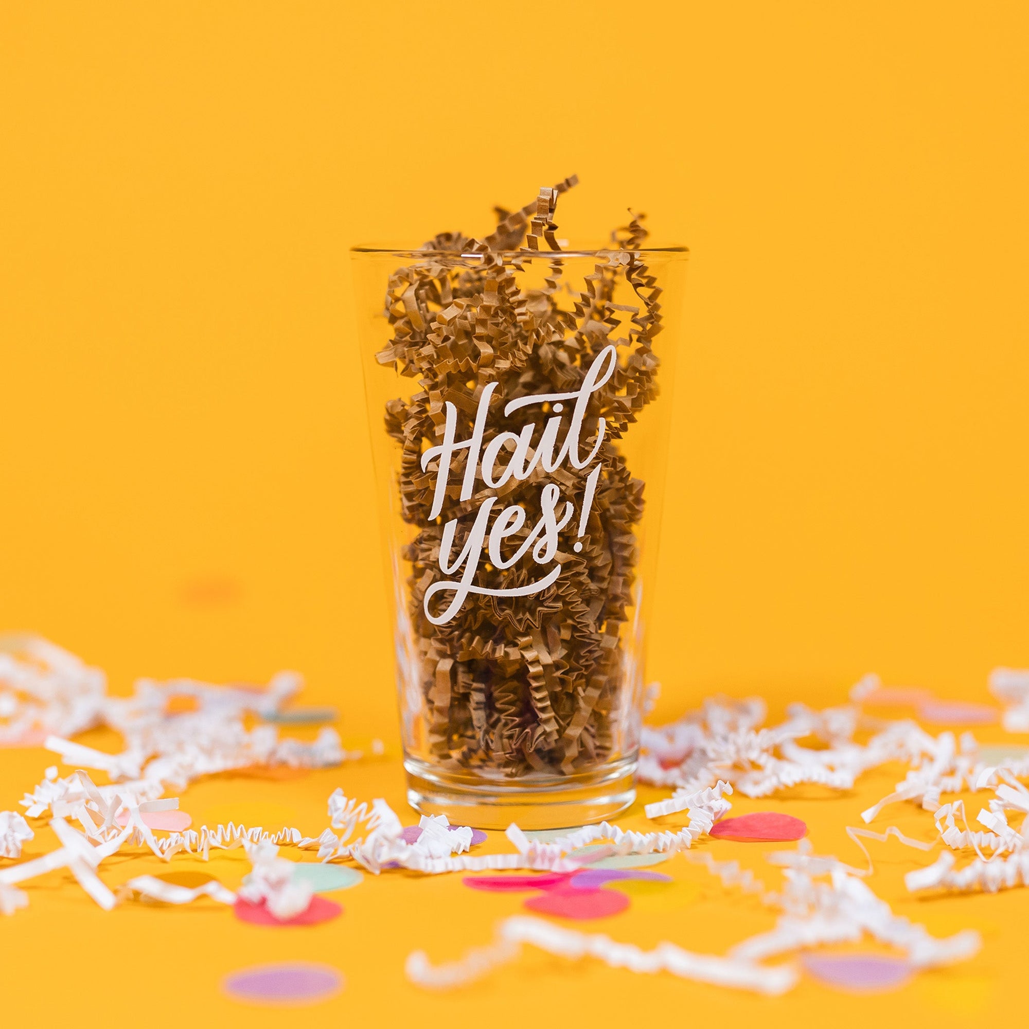 On a sunny mustard background sits a pint glass with white crinkle and big, colorful confetti scattered around. This beer pint glass is filled with kraft crinkle and it says in white hand lettering "Hail Yes!"