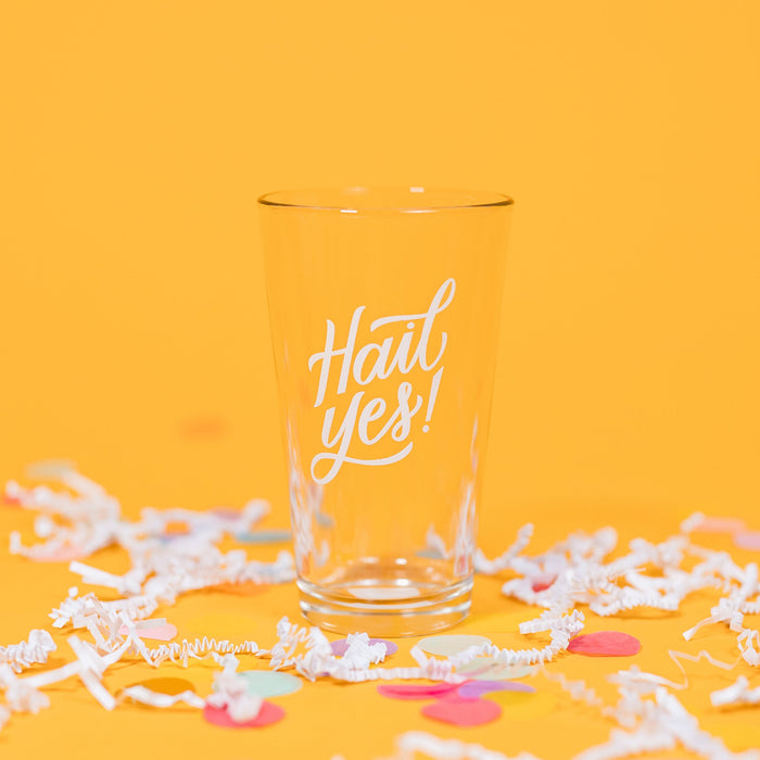 On a sunny mustard background sits a pint glass with white crinkle and big, colorful confetti scattered around. This beer pint glass says in white hand lettering "Hail Yes!"