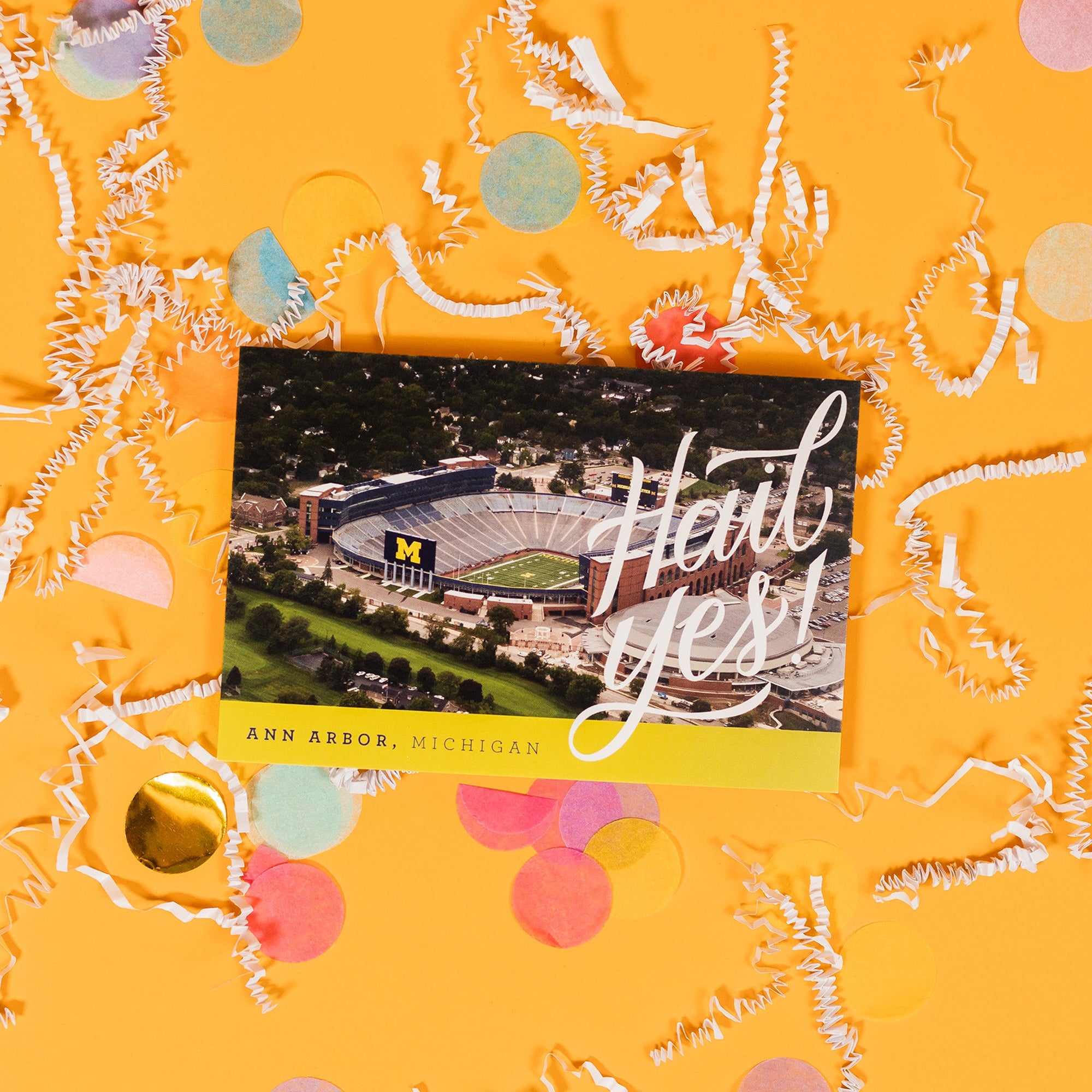 On a sunny mustard background sits a postcard with white crinkle and big, colorful confetti scattered around. This postcard has a picture of The Big House stadium with a yellow stripe at the bottom. It says in white hand-lettering "Hail Yes!" and in the yellow stripe it says "ANN ARBOR, MICHIGAN."