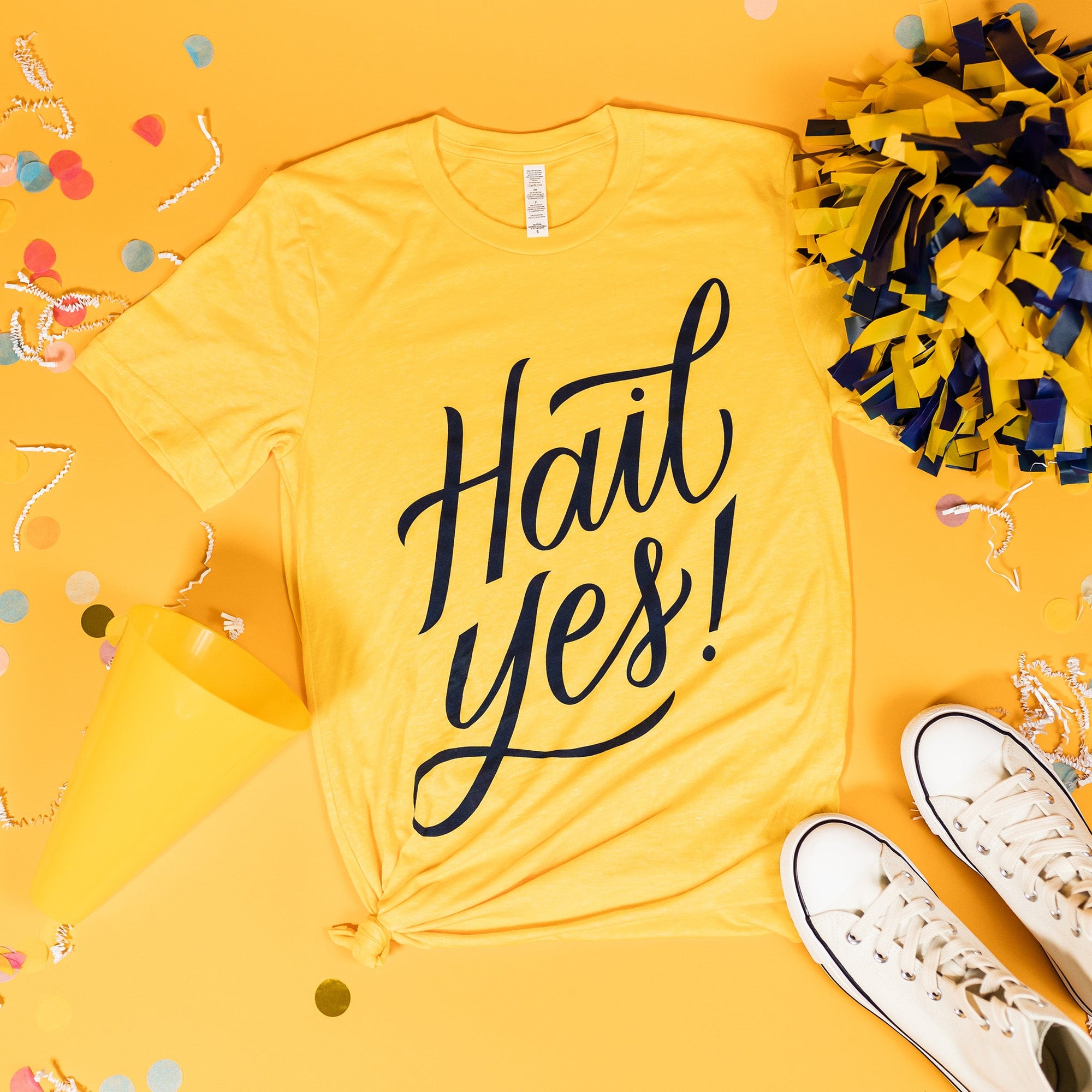 On a sunny mustard background sits a t-shirt with white crinkle and big, colorful confetti scattered around. There is a yellow and navy pom pom, a yellow mini megaphone, and a pair of white sneakers. This yellow tee says "Hail Yes!" in blue lettering.