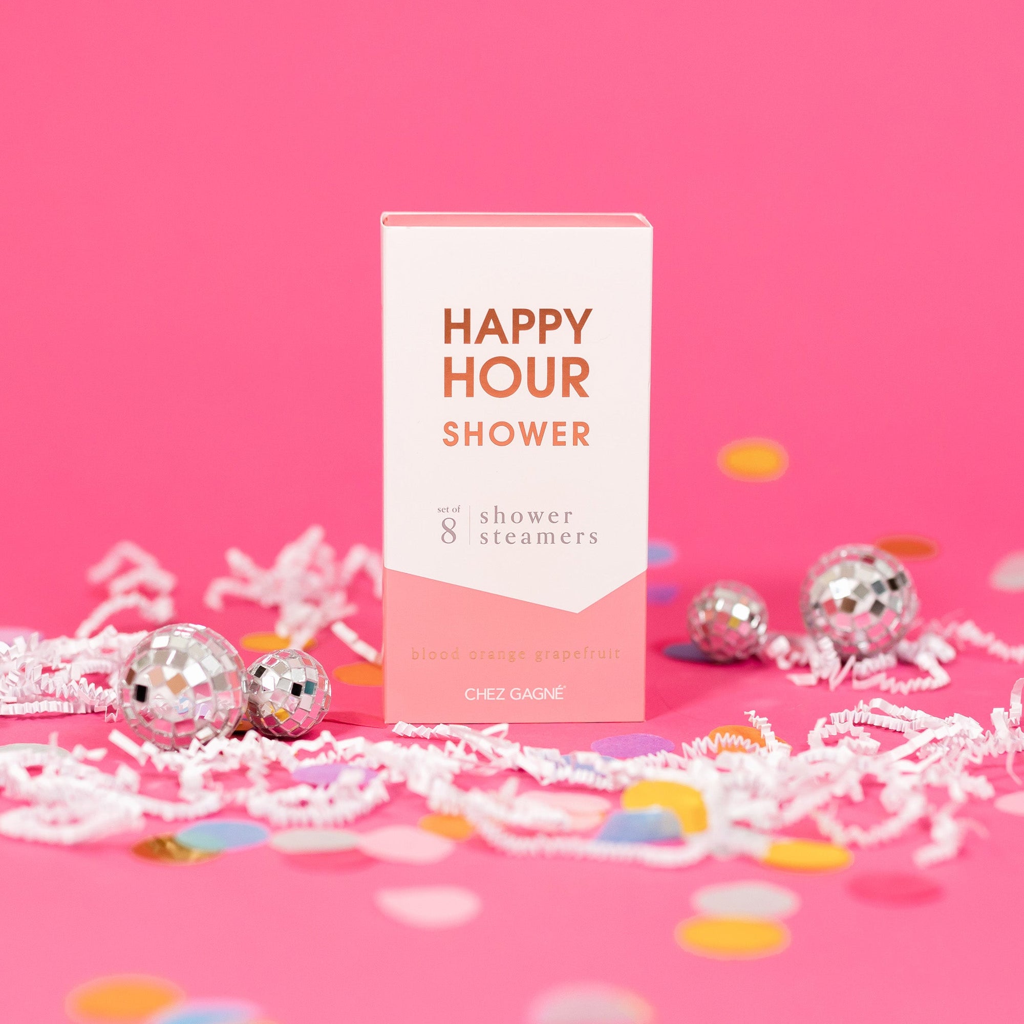 On a hot pink background sits a box with white crinkle and big, colorful confetti scattered around. There are mini disco balls. This picture is a close-up of a white and coral package that says "HAPPY HOUR SHOWER" in gold foil, all caps block lettering. Under it says "set of 8" and " shower steamers" in grey, lowercase serif font. At the bottom it says "blood orange grapefruit" in gold foil, lower case serif font. 