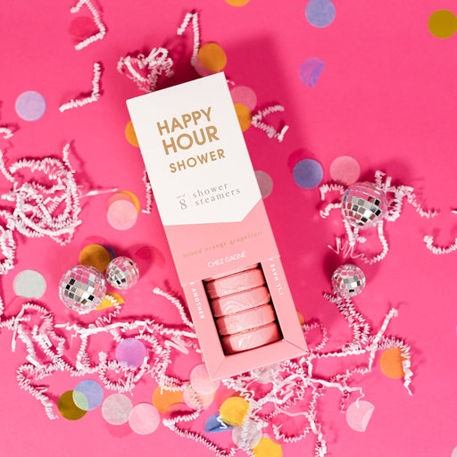 On a hot pink background sits an opened box with white crinkle and big, colorful confetti scattered around. There are mini disco balls. This picture is a close-up of a white and coral package that says "HAPPY HOUR SHOWER" in gold foil, all caps block lettering. Under it says "set of 8" and " shower steamers" in grey, lowercase serif font. At the bottom it says "blood orange grapefruit" in gold foil, lower case serif font. The box is opened to reveal the pink shower steamers in it.