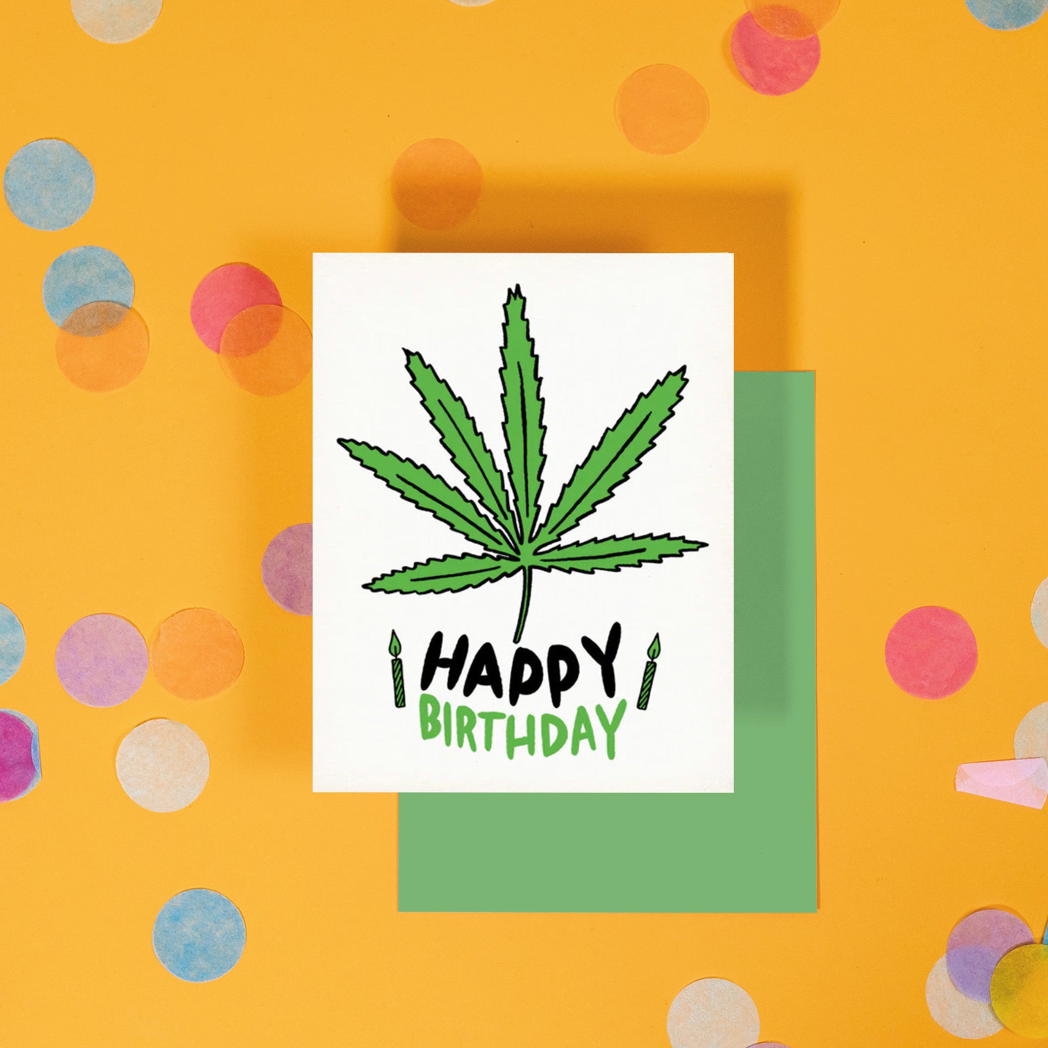 On a sunny mustard background is a greeting card and envelope with big, colorful confetti scattered around. The white greeting card has an illustration of a green and black marijuana leaf and candles and it says "HAPPY BIRTHDAY" in black and green, handwritten lettering. The kelly green envelope sits under the card.