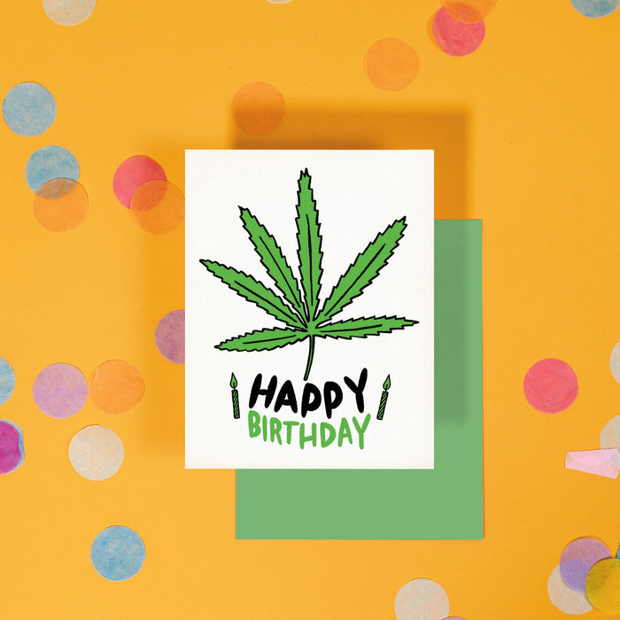On a sunny mustard background is a greeting card and envelope with big, colorful confetti scattered around. The white greeting card has an illustration of a green and black marijuana leaf and candles and it says "HAPPY BIRTHDAY" in black and green, handwritten lettering. The kelly green envelope sits under the card.