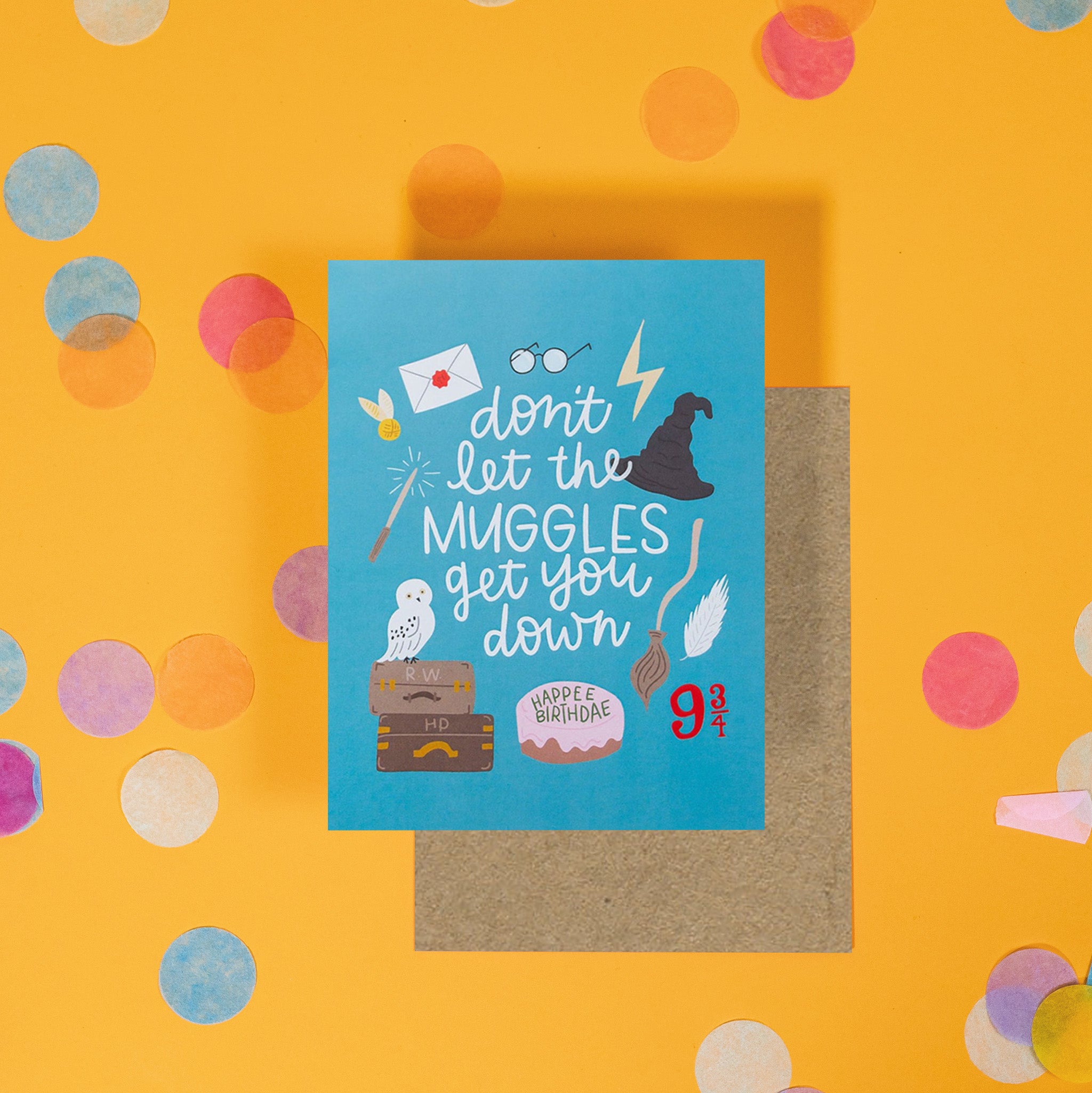 On a sunny mustard background is a greeting card and envelope with big, colorful confetti scattered around. The Harry Potter inspired greeting card has a pacific blue background with illustrations of icons from Harry Potter. It says "don't let the muggles get you down" in a white, handwritten script lettering and a birthday cake at the bottom that says "HAPPEE BIRTHDAE" in a handwritten lettering. The kraft envelope sits under the card.