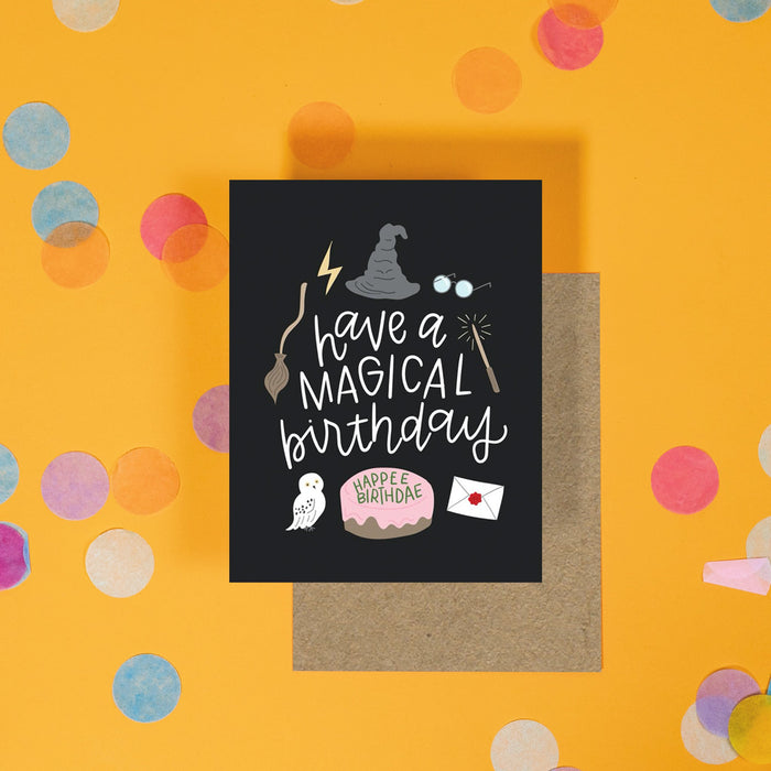 On a sunny mustard background is a greeting card and envelope with big, colorful confetti scattered around. The Harry Potter inspired greeting card has a black background with illustrations of icons from Harry Potter. It says "have a magical birthday" in a white, handwritten script lettering and a birthday cake at the bottom that says "HAPPEE BIRTHDAE" in a handwritten lettering. The kraft envelope sits under the card.