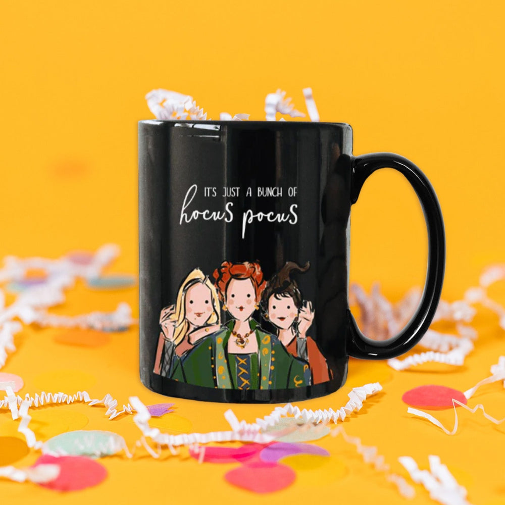 On a sunny mustard background sits a mug with white crinkle and big, colorful confetti scattered around. This black mug has a colorfuI illustration of three women form the hit movie 'Hocu Pocus'. It says "IT'S JUST A BUNCH OF hocus pocus" in white, handwritten lettering. There is white crinkle in the mug.