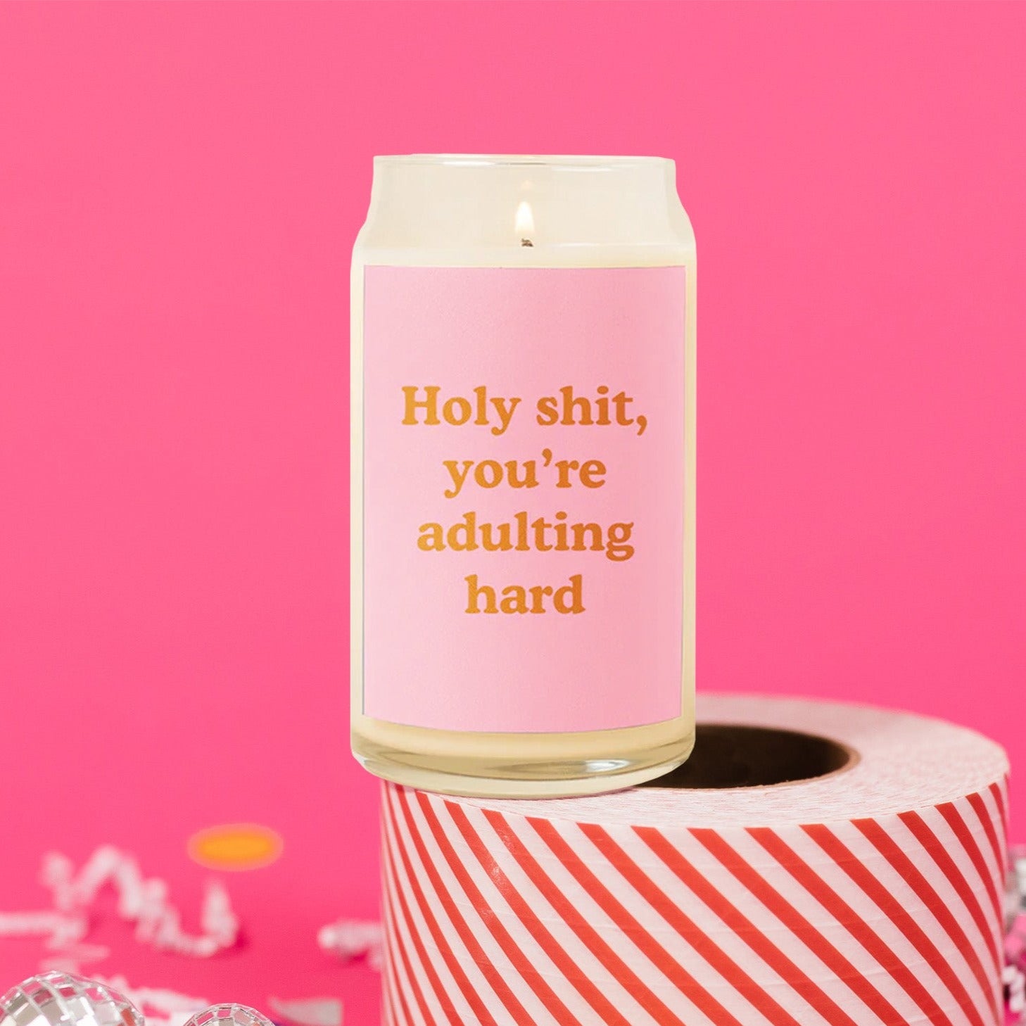 On a hot pink background sits a candle atop a roll of red and white striped packing tape. There are white crinkle and big, colorful confetti scattered around.. This beer can inspired glass candle is lit and has a bubblegum pink label on the front. It says "Holy shit, you're adulting hard" in a dark orange, thick serif font.