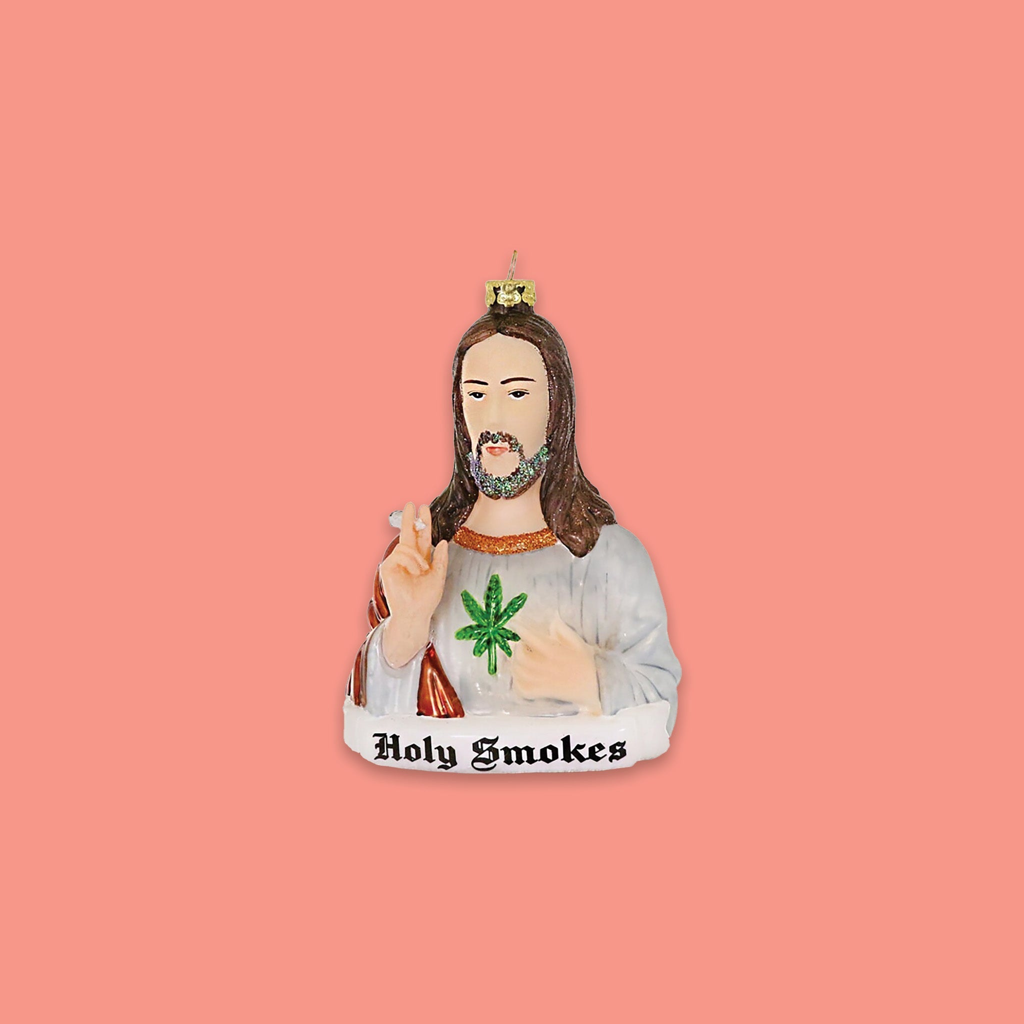 On a coral pink background sits an ornament. This glass ornament is a bust of Jesus holding a marijuana joint and there is a cannabis leaf on the shirt. At the bottom it says "Holy Smokes" in black, olde english lettering.