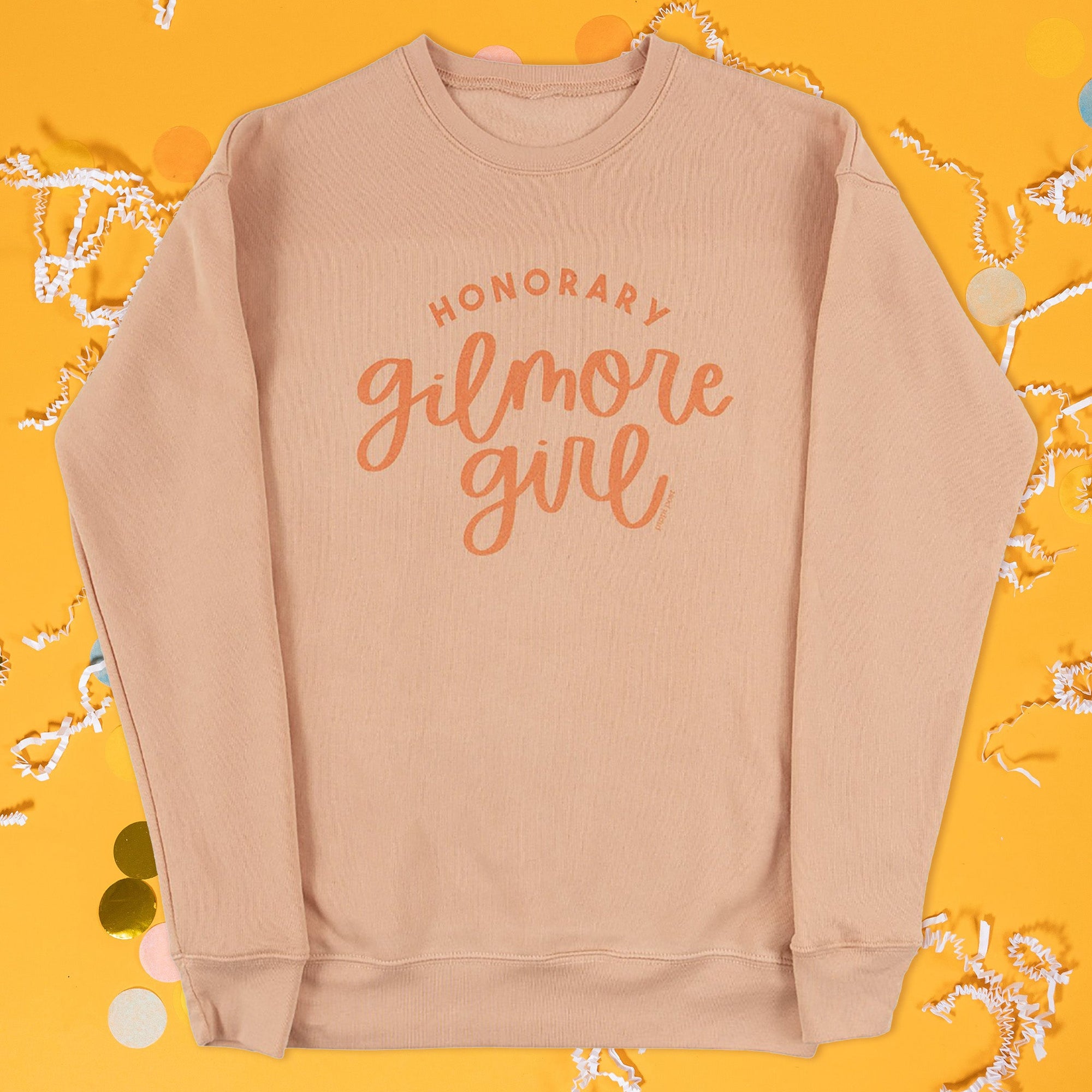 On a sunny mustard background lays a sweatshirt with white crinkle and big, colorful confetti scattered around. The sweatshirt is a pretty peach color that says "Honorary Gilmore Girl" in orange. The word "Honorary" is in an all caps, sans serif font and the words "Gilmore Girls" is in a hand drawn script font.