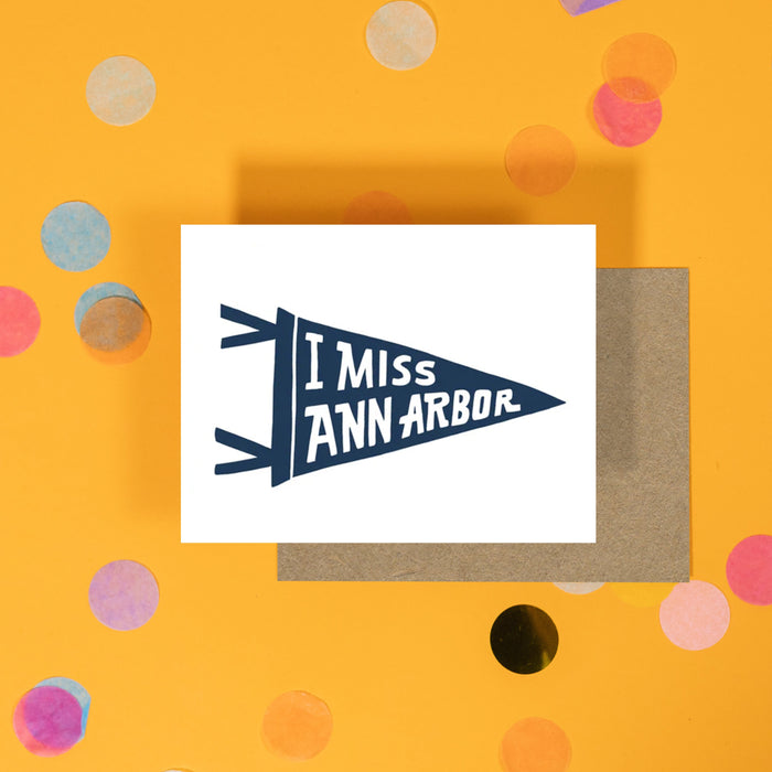 On a sunny mustard background sits a card and envelope with white crinkle and big, colorful confetti scattered around. This white card has a vintage navy pennant that says "I MISS ANN ARBOR" in white, all caps handwritten lettering.