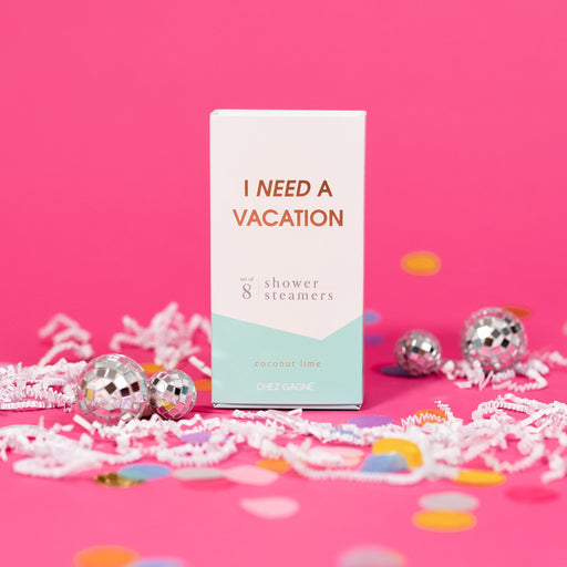 On a hot pink background sits a box with white crinkle and big, colorful confetti scattered around. There are mini disco balls. This picture is a close-up of a white and pool blue package that says "I NEED A VACATION" in gold foil, all caps block lettering. Under it says "set of 8" and " shower steamers" in grey, lowercase serif font. At the bottom it says "coconut lime" in gold foil, lower case serif font.