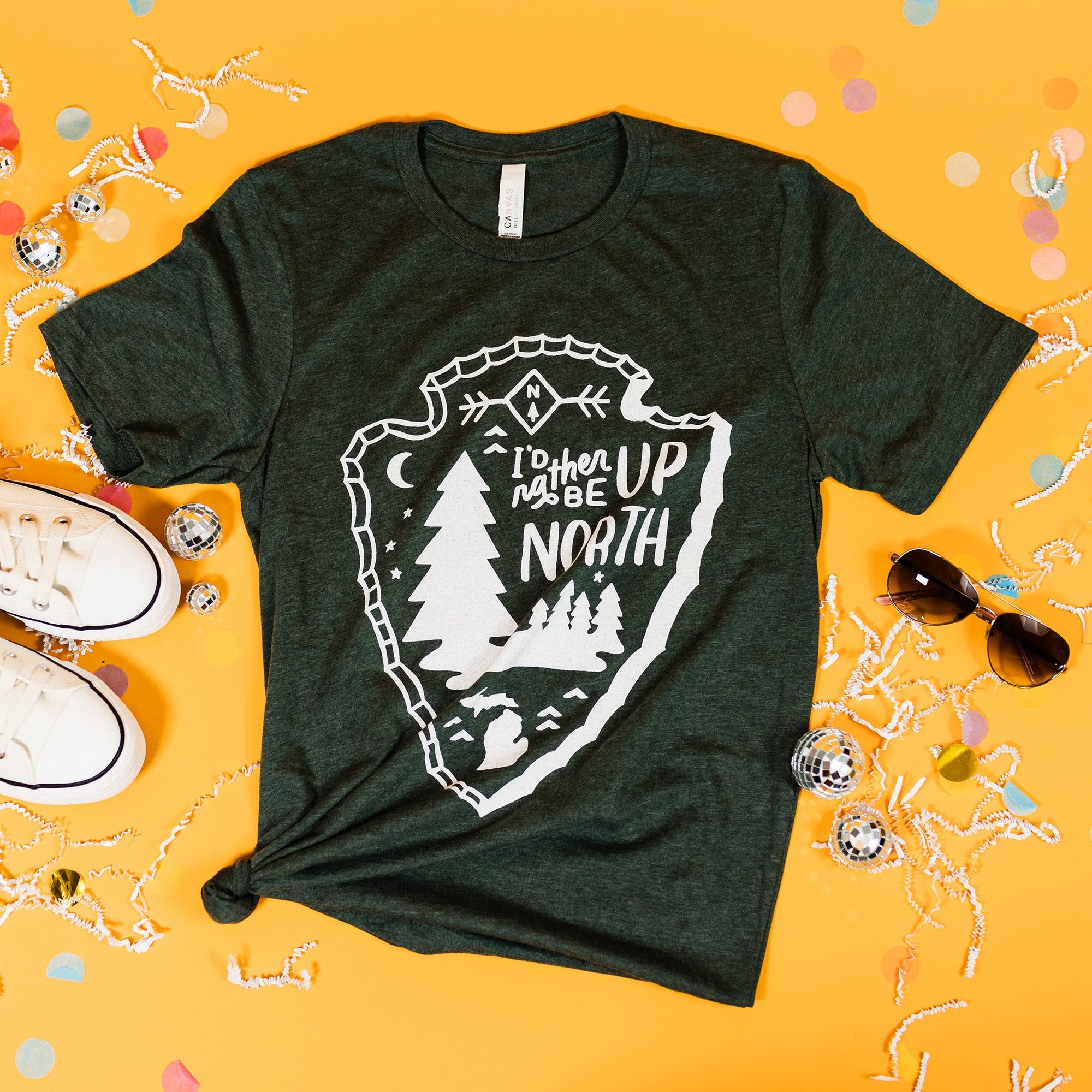 On a sunny mustard background sits a t-shirt with white crinkle and big, colorful confetti scattered around. There are mini disco balls, sunglasses, and a pair of white sneakers. This weathered forest green tee features hand-lettering and various custom illustrations enclosed by an arrowhead design in white and says "I'd rather be UP NORTH."