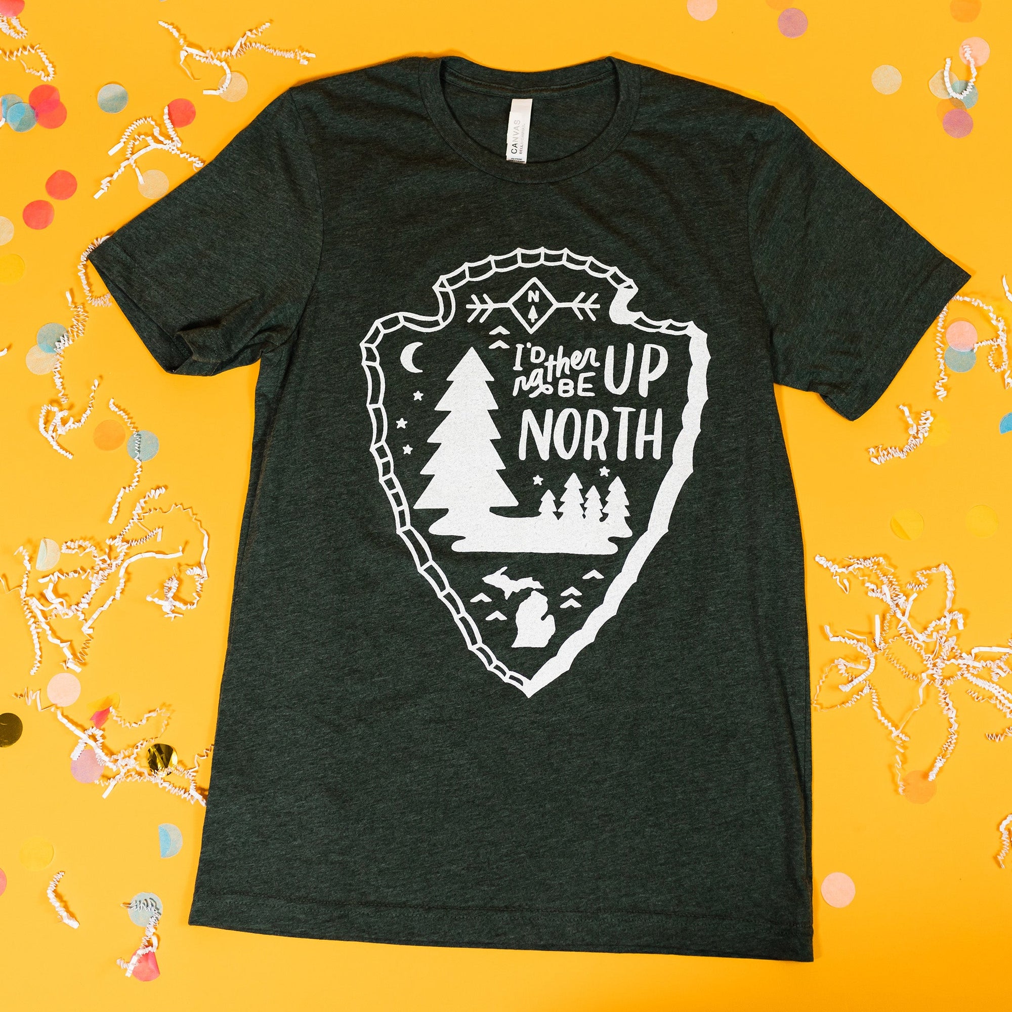 On a sunny mustard background sits a t-shirt with white crinkle and big, colorful confetti scattered around. This weathered forest green tee features hand-lettering and various custom illustrations enclosed by an arrowhead design in white and says "I'd rather be UP NORTH."