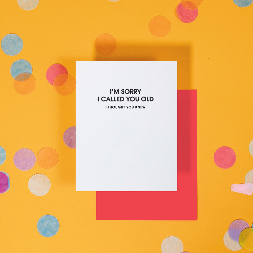 On a sunny mustard background is a greeting card and envelope with big, colorful confetti scattered around. The white greeting card says "I'M SORRY I CALLED YOU OLD" in a black, all caps block font at the top. Under it in a smaller size black, all caps font it says "I THOUGHT YOU KNEW." A red envelope sits under the card.