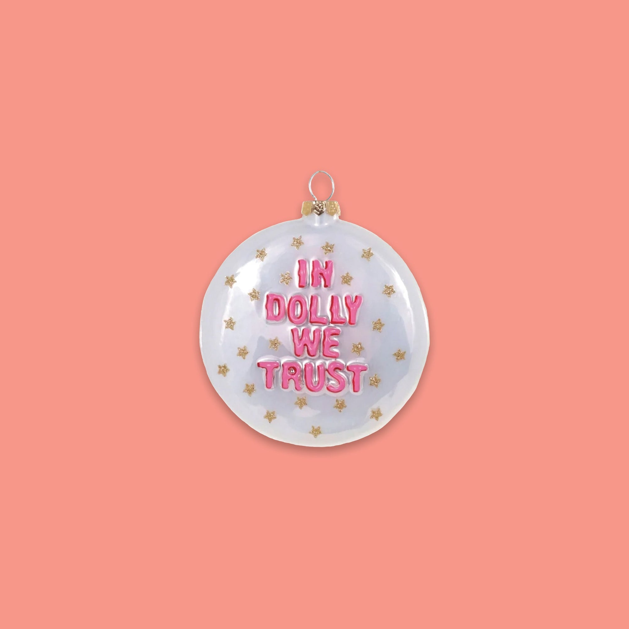 On a coral pink background sits a round ornament. This is a white ornament that has gold glitter stars and in pink it says "IN DOLLY WE TRUST."