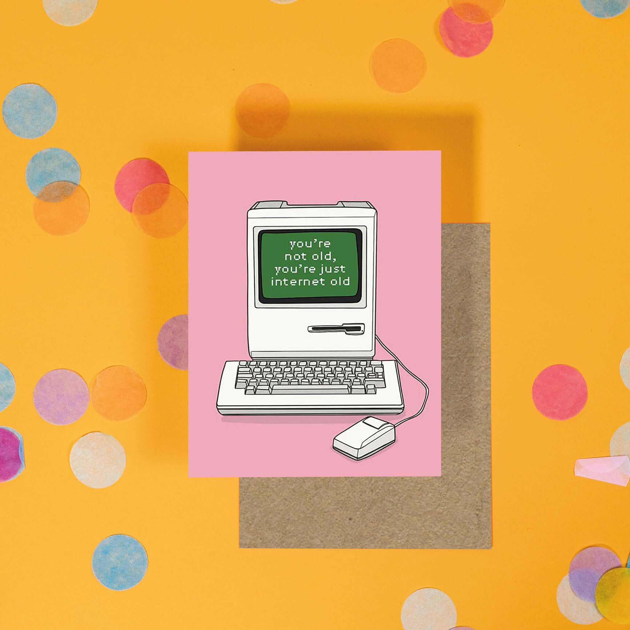 On a sunny mustard background is a greeting card and envelope with big, colorful confetti scattered around. The bubblegum pink greeting card has an illustration of an old computer monitor and keyboard with mouse that says "you're not old, you're just internet old" in a computer style font. A kraft envelope sits under the card.