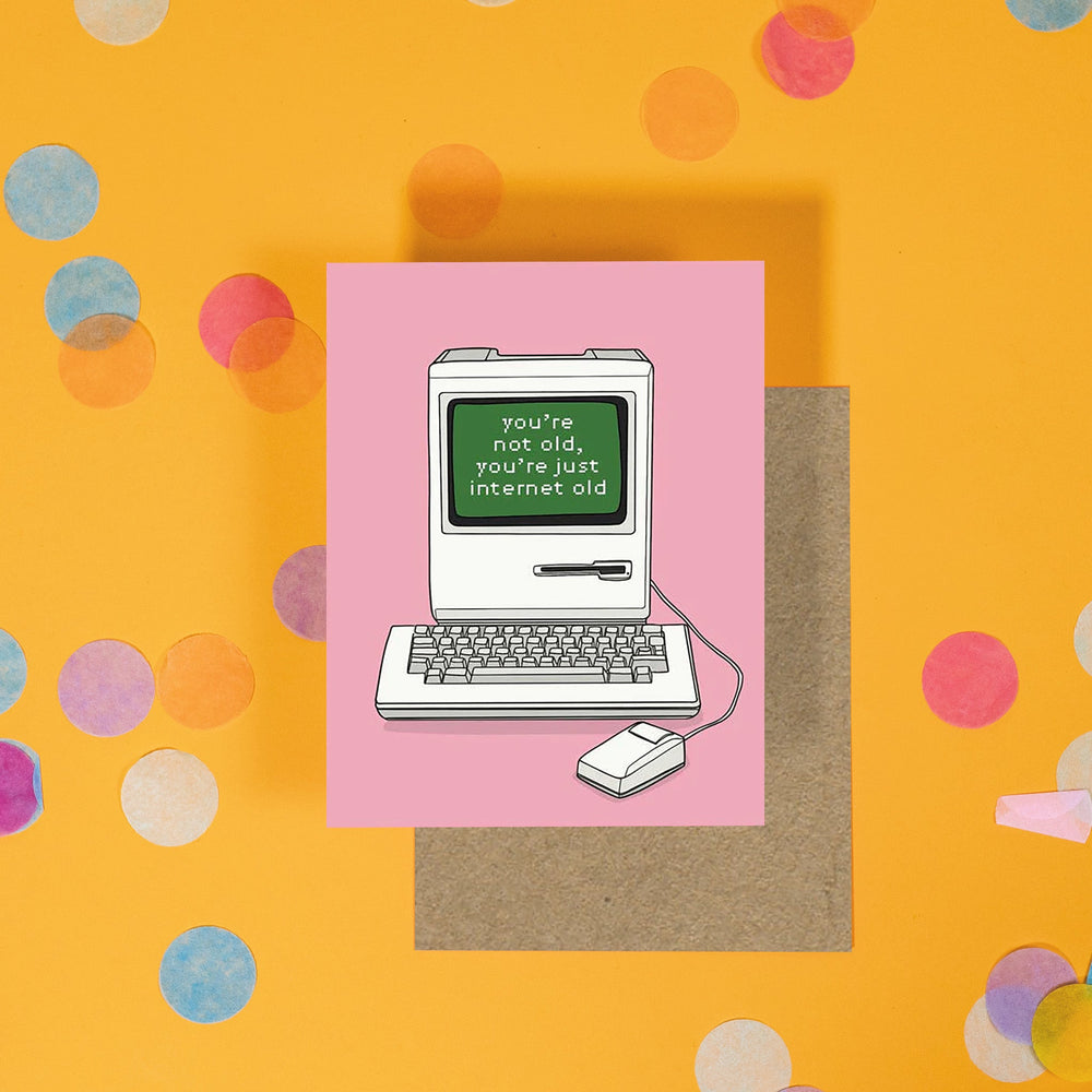 On a sunny mustard background is a greeting card and envelope with big, colorful confetti scattered around. The bubblegum pink greeting card has an illustration of an old computer monitor and keyboard with mouse that says "you're not old, you're just internet old" in a computer style font. A kraft envelope sits under the card.