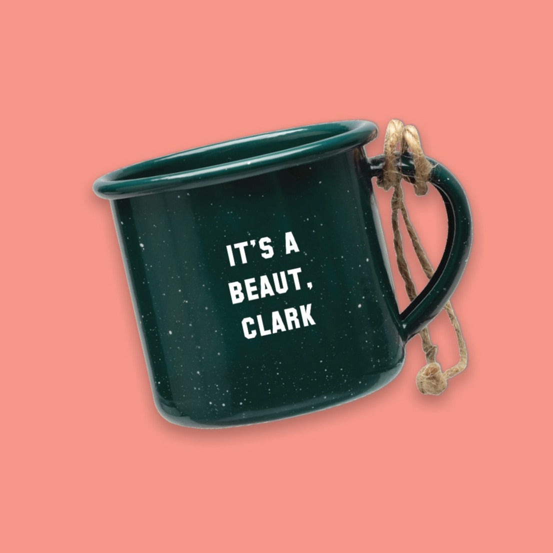 On a coral pink background sits a dark green mini campfire mug with white specks. It says "IT'S A BEAUT, CLARK" in white collegiate lettering. It has twine on the handle.