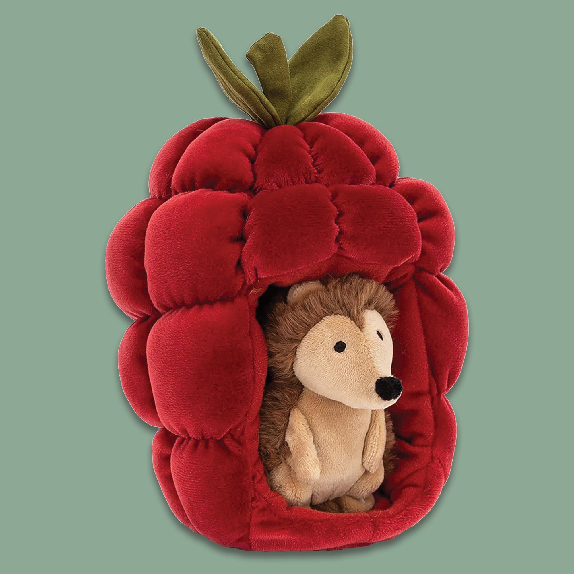 On a moss green background sits a Jellycat Brambling Hedgehog inside of a red raspberry. It is squishy in stretchy lined scarlet, wiht pockets of softness and funky green leaves. The butterscotch hoglet has a snuffly nose and soft cocoa spines.