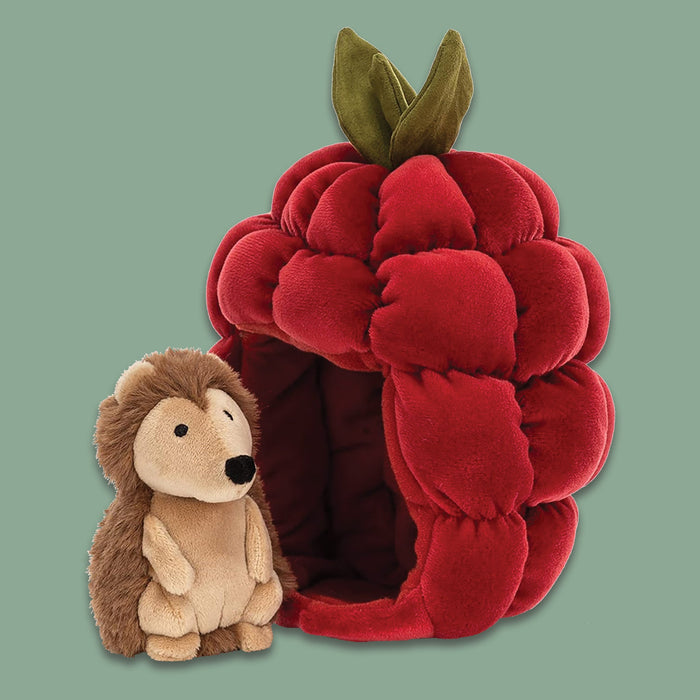 On a moss green background sits a Jellycat Brambling Hedgehog beside a red raspberry. The raspberry is squishy in stretchy lined scarlet, wiht pockets of softness and funky green leaves. The butterscotch hoglet has a snuffly nose and soft cocoa spines.
