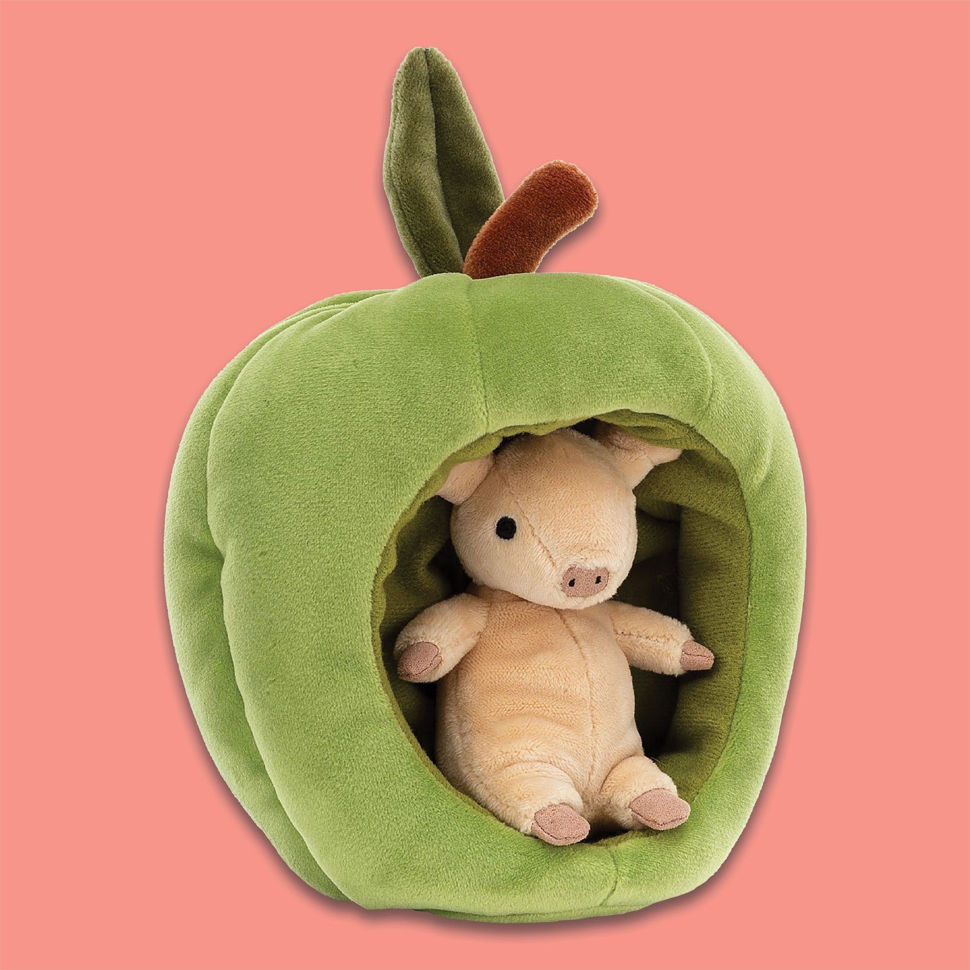 On a coral backgrounds sits a Jellycat Brambling Pig inside a green apple. This apple is stretchy-soft and cozily lined, with a velvety leaf on top. The pig is peach and has the softest fur.