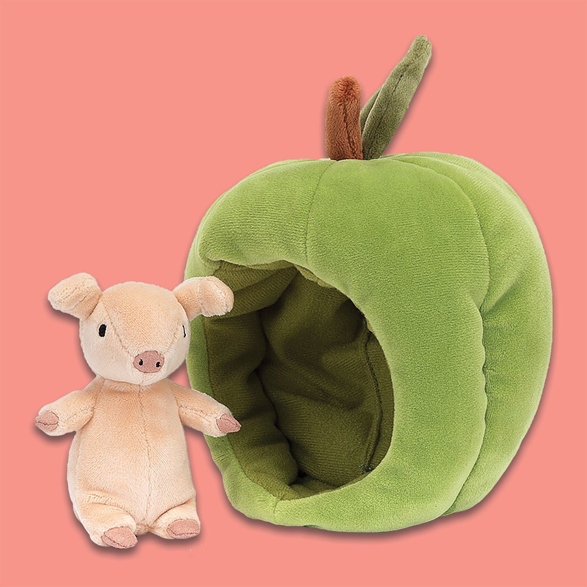 On a coral backgrounds sits a Jellycat Brambling Pig beside a green apple. This apple is stretchy-soft and cozily lined, with a velvety leaf on top. The pig is peach and has the softest fur.