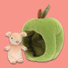 On a coral backgrounds sits a Jellycat Brambling Pig beside a green apple. This apple is stretchy-soft and cozily lined, with a velvety leaf on top. The pig is peach and has the softest fur.