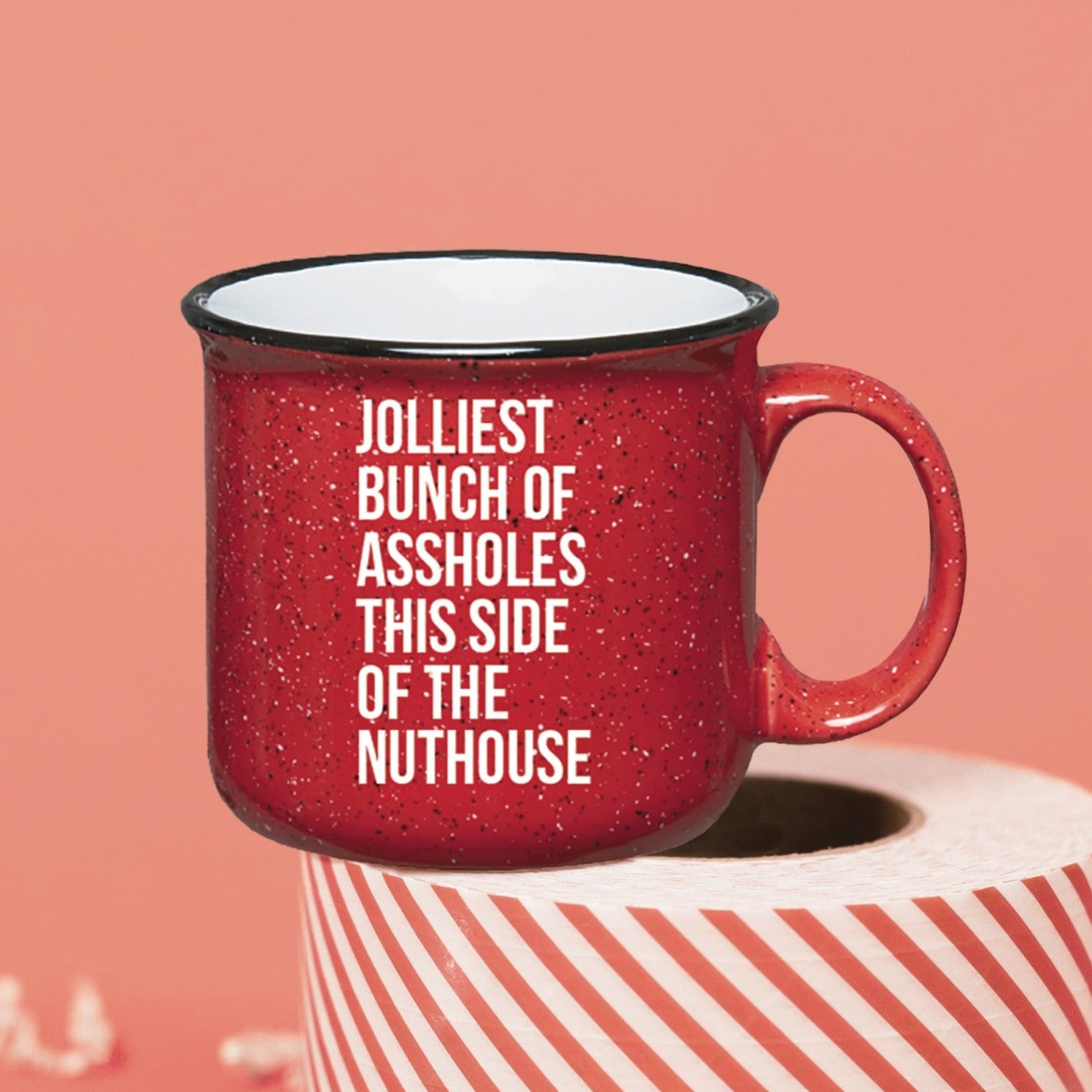 On a pink coral background sits a campfire mug on a roll of red and white packing tape. There is white crinkle. This Christmas Vacation inspired mug is red with black and white specks and it's say in white lettering "JOLLIEST BUNCH OF ASSHOLES THIS SIDE OF THE NUTHOUSE."