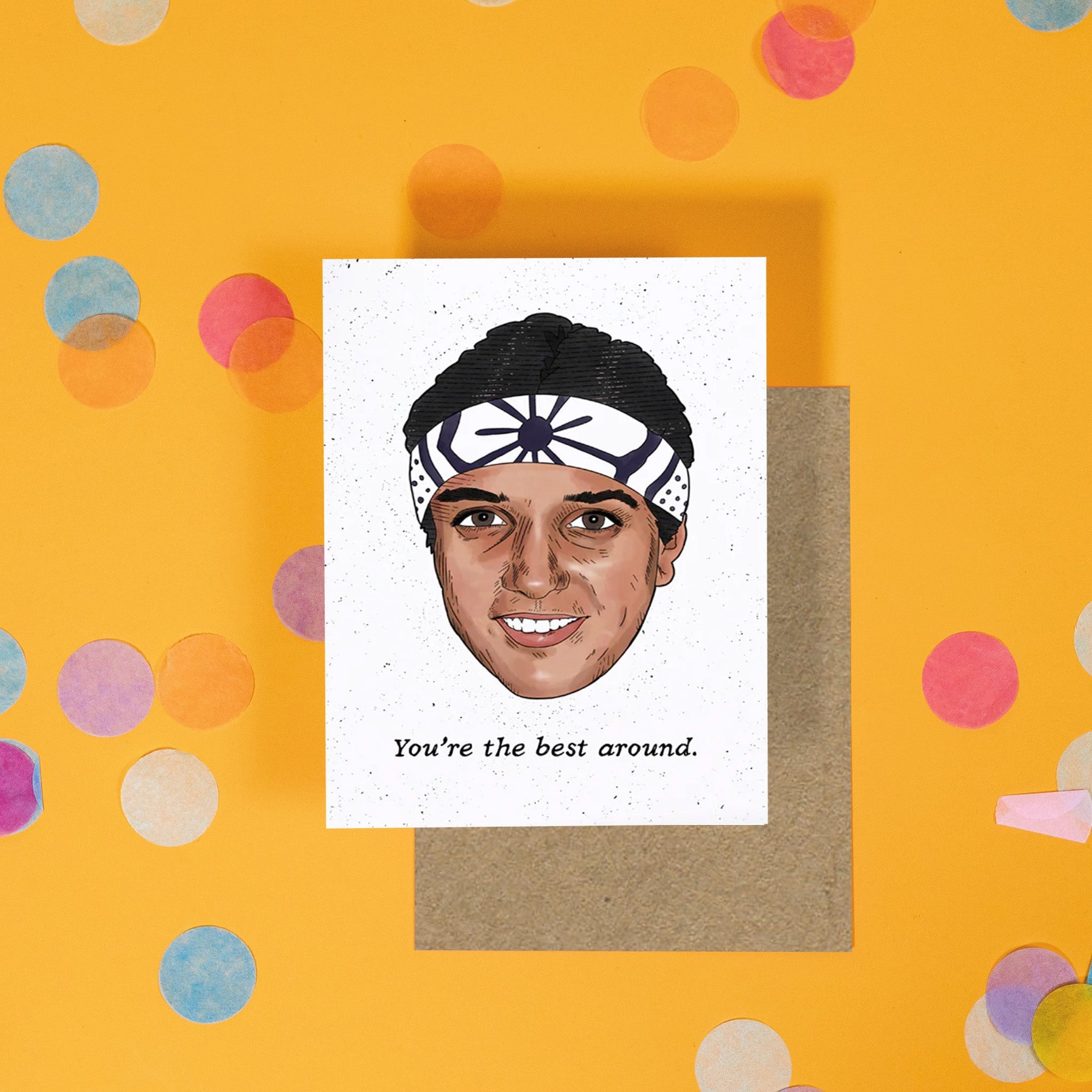 On a sunny mustard background is a greeting card and envelope with big, colorful confetti scattered around. The Karate Kid inspired white greeting card with black specks has an illustration of the Karate Kid with a black and white japanese style bandana on his head. Under it says "You're the best around." in a black handwritten lettering font. A kraft envelope sits under the card.