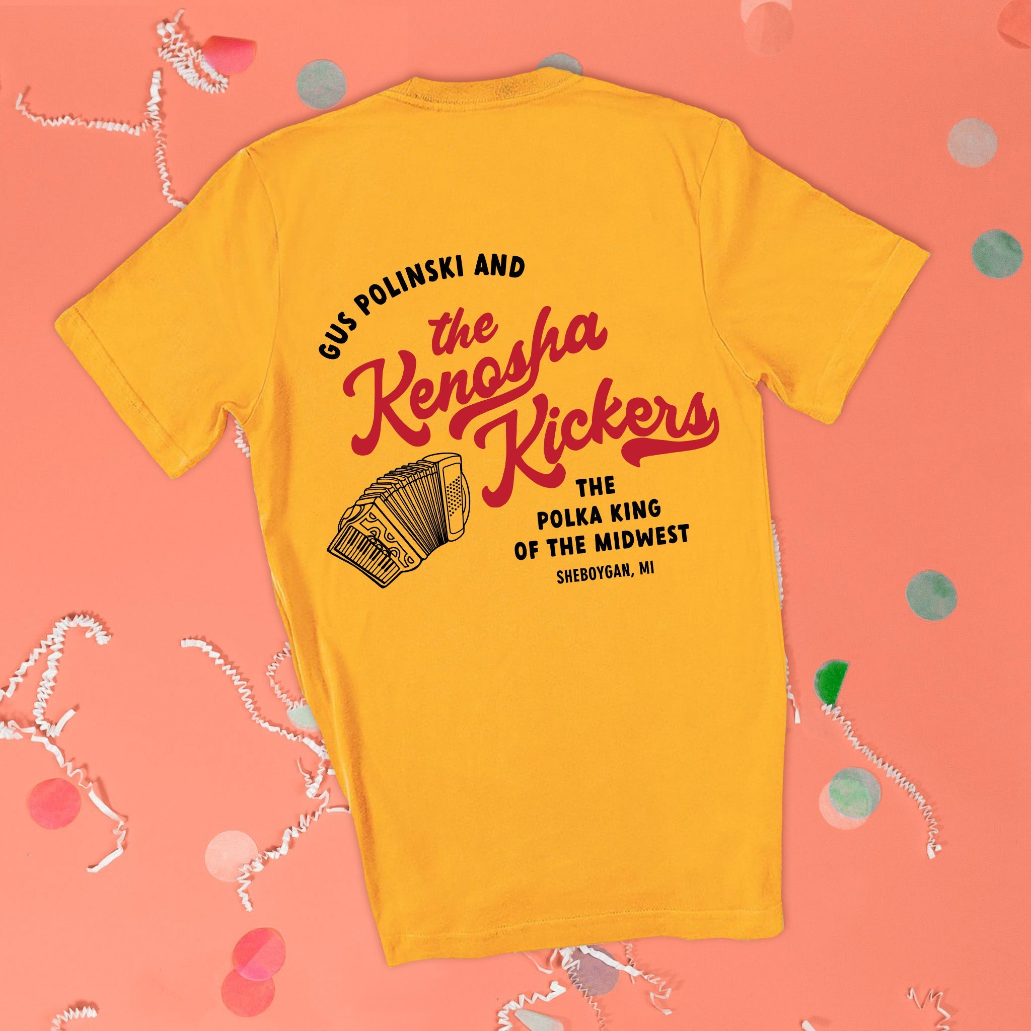 On a pink coral background sits the back of a t-shirt with white crinkle and big, colorful confetti scattered around. This Home Alone Inspired crewneck sunny mustard t-shirt has red script lettering black lettering. There is an illustration of an accordian and it says "GUS POLINSKI AND the Kenosha Kickers THE POLKA KIN OF THE MIDWEST SHEBOYGAN, MI."