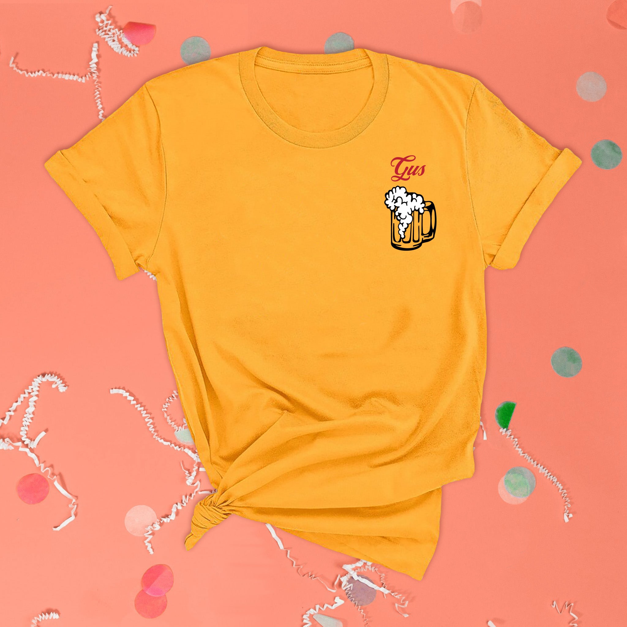 On a pink coral background sits the front of a t-shirt with white crinkle and big, colorful confetti scattered around. This Home Alone Inspired crewneck sunny mustard t-shirt has red script lettering and and illustration of a beer mug with beer foam. It says "Gus."