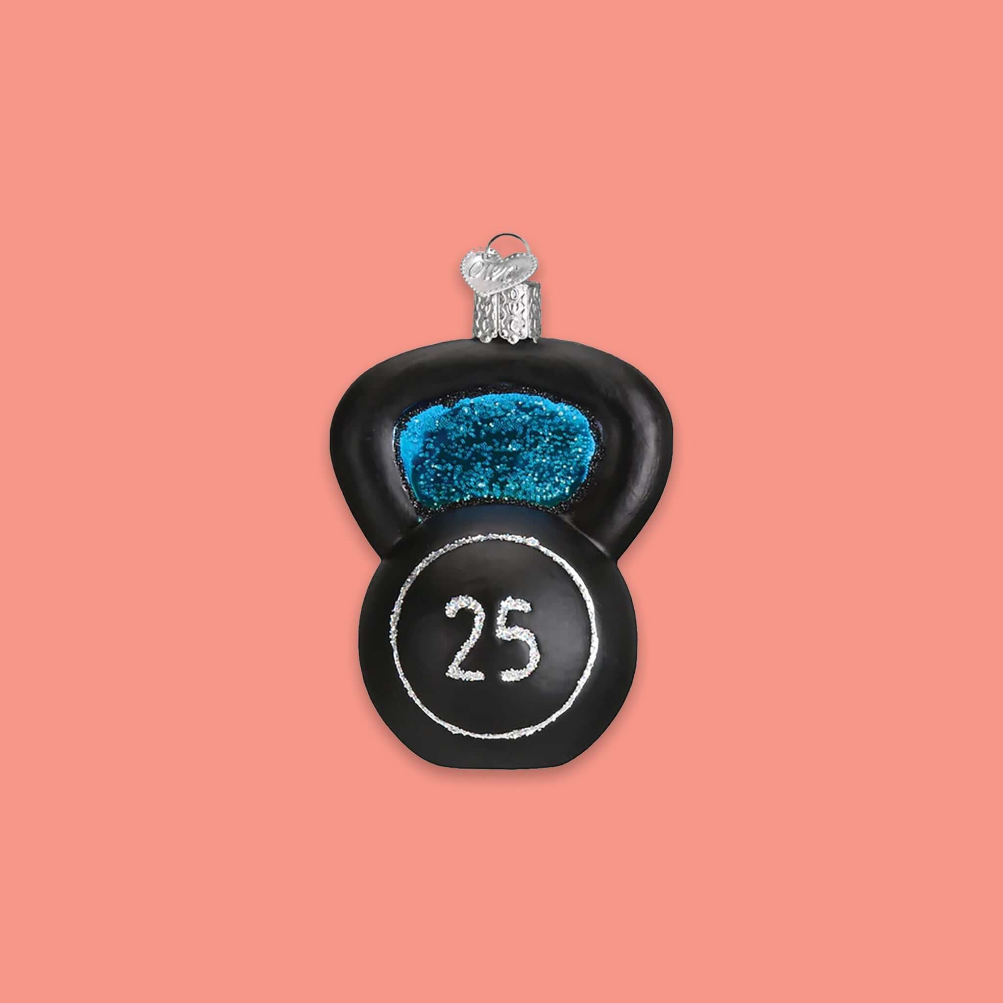 On a coral pink background sits an ornament. This glass ornament is of a black kettlebell with a silver glitter circle and the number '25' in silver glitter inside the circle. 