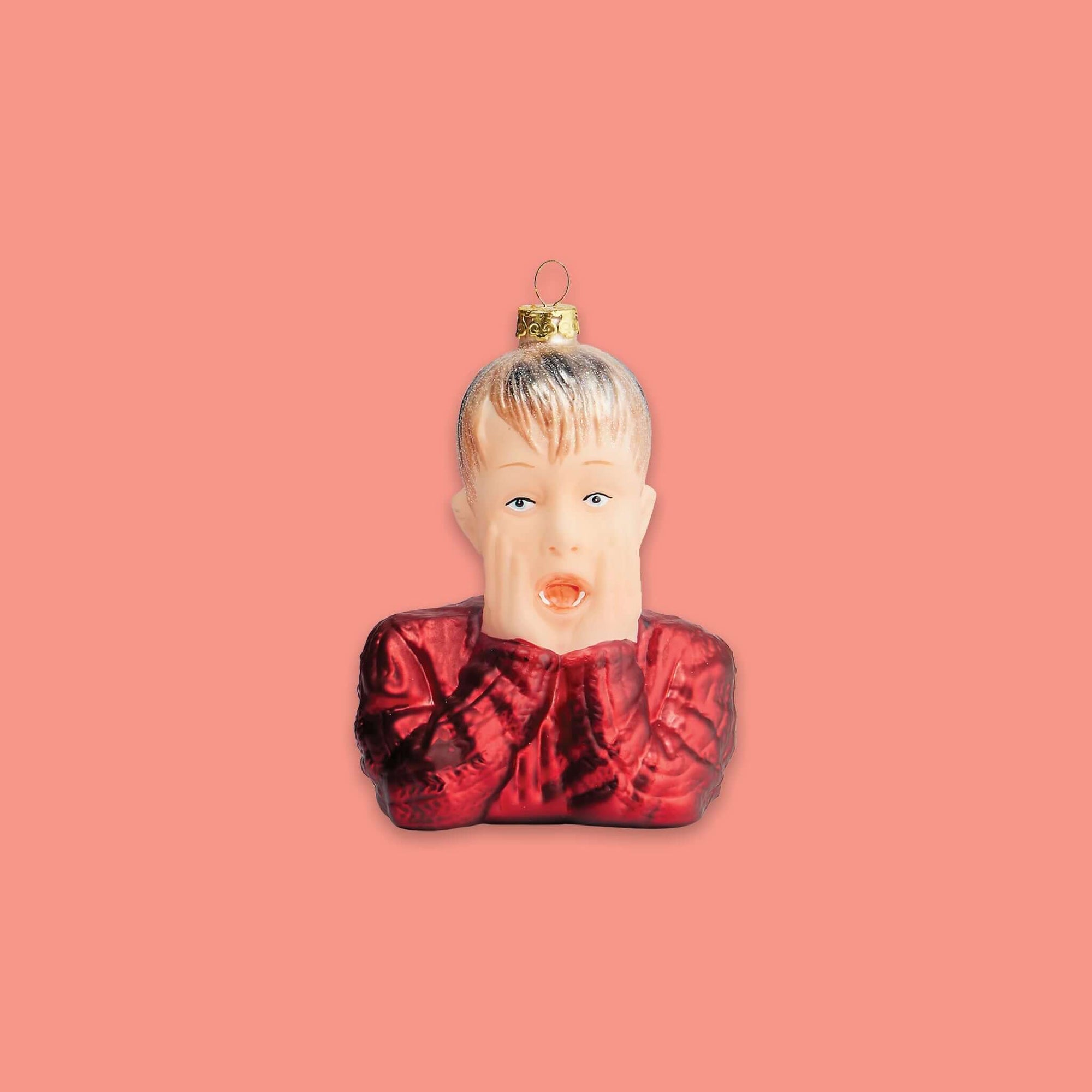 On a coral pink background sits an ornament. This glass Home Alone inspired ornament is a bust of Kevin McAllister wearing a red sweater and looking surprised with his hands on his cheeks.